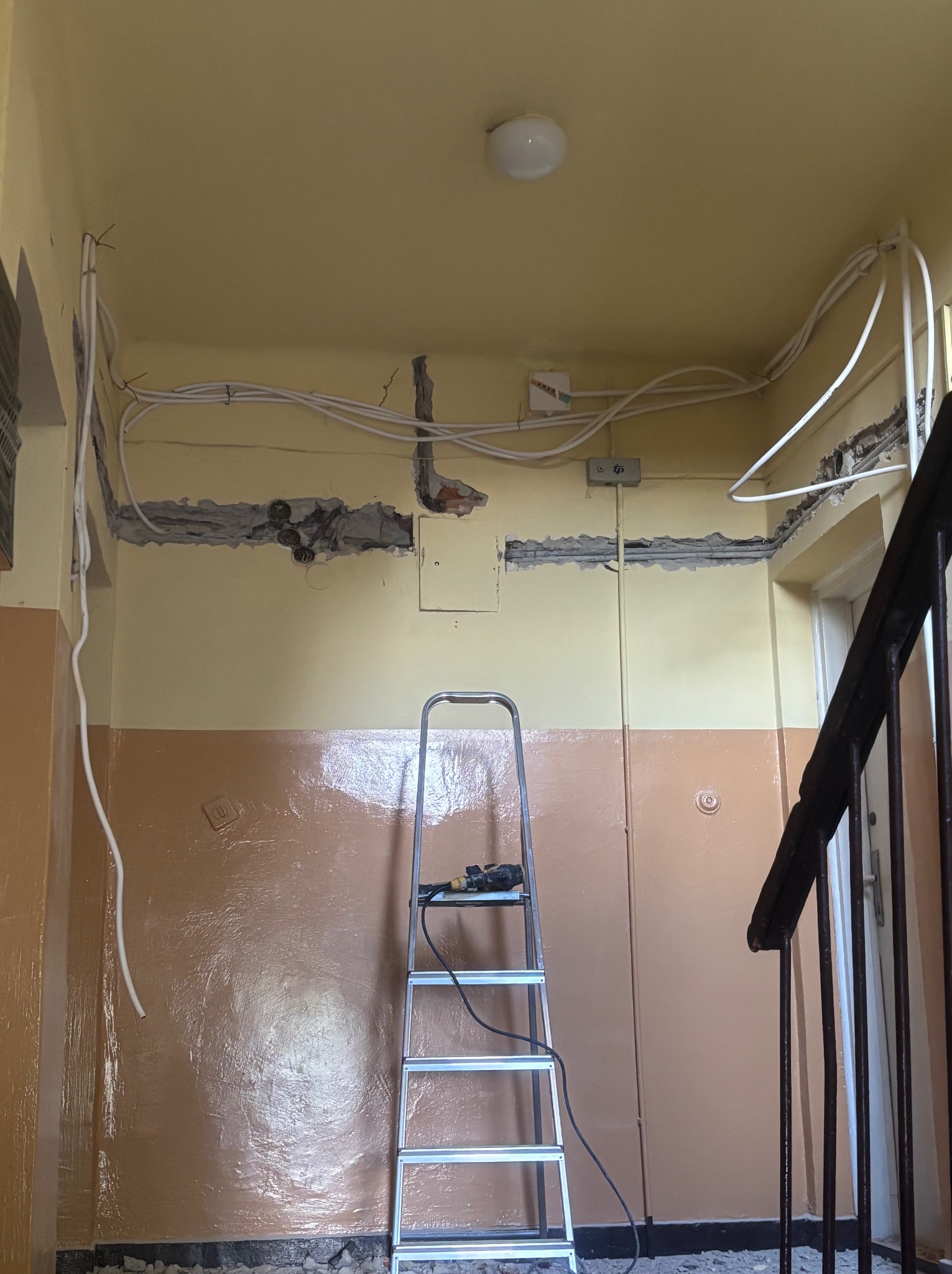 Klatka schodowa w trakcie remontu elektrycznego: widoczne kable w bruzdach, drabina z narzędziami. Ściany dwukolorowe, oświetlenie sufitowe. Ujęcie z dołu.