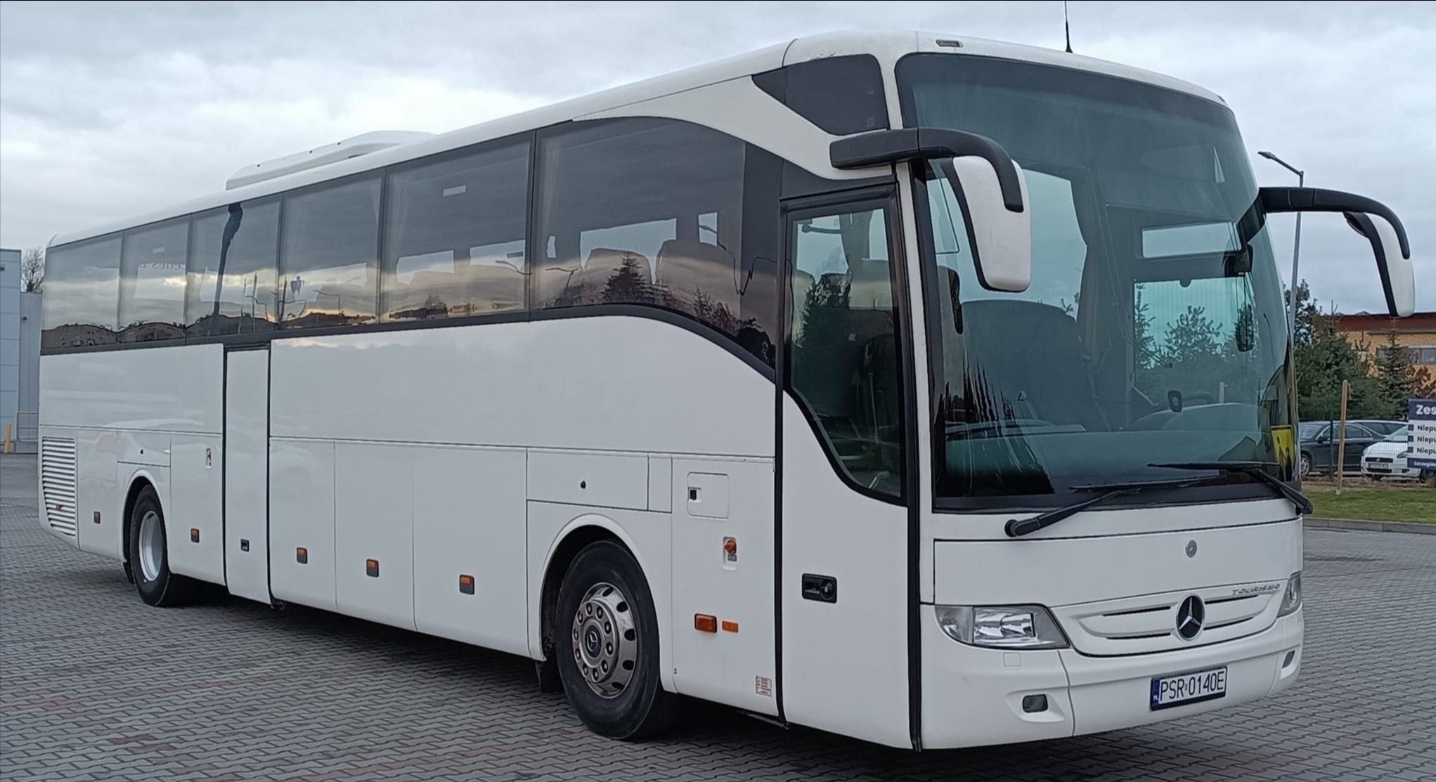 Biały autokar Mercedes-Benz Tourismo z widoczną tablicą rejestracyjną PSR 0140E, zaparkowany na kostce brukowej, z budynkami i zielenią w tle.