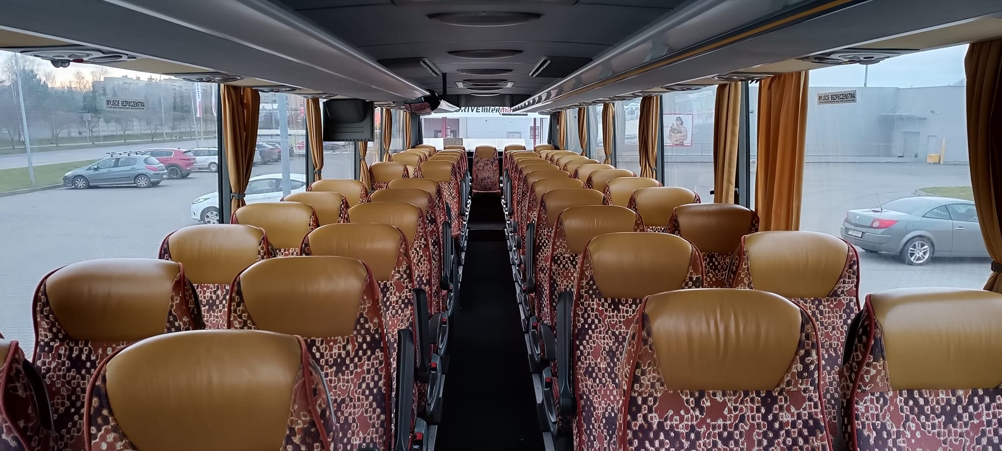 Wnętrze autobusu z rzędami foteli z brązową tapicerką i wzorzystymi zagłówkami. Widok na okna z zasłonami i parking z samochodami na zewnątrz.
