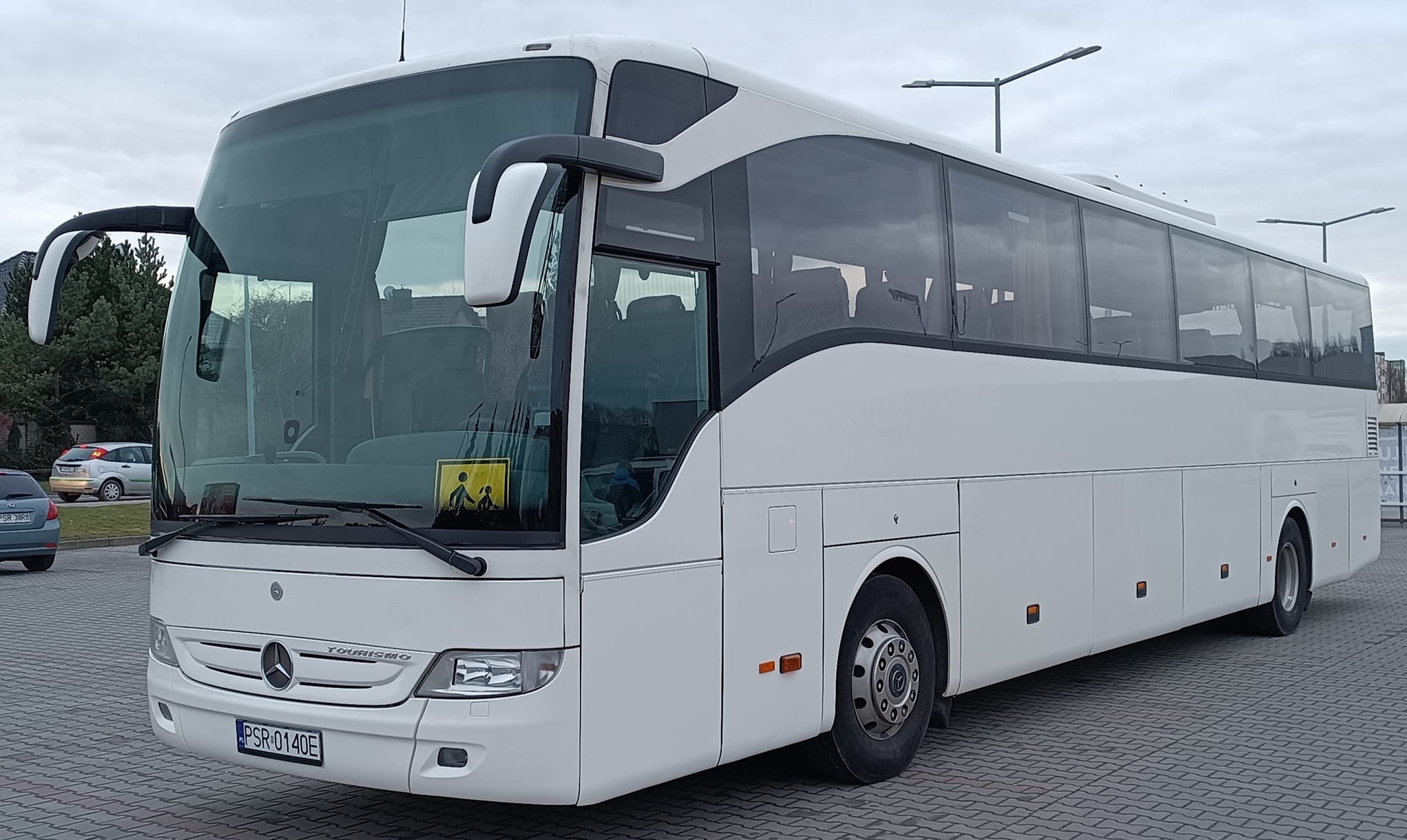 Biały autokar Mercedes-Benz Tourismo z żółtą naklejką 'dzieci' na szybie, zaparkowany na kostce brukowej. Widok z przodu i boku, pochmurne niebo w tle.
