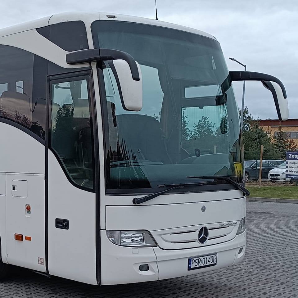 Biały autokar Mercedes-Benz Tourismo z widocznym przodem i szybą, zaparkowany na placu z kostki brukowej. Widoczne lusterka i tablica rejestracyjna.