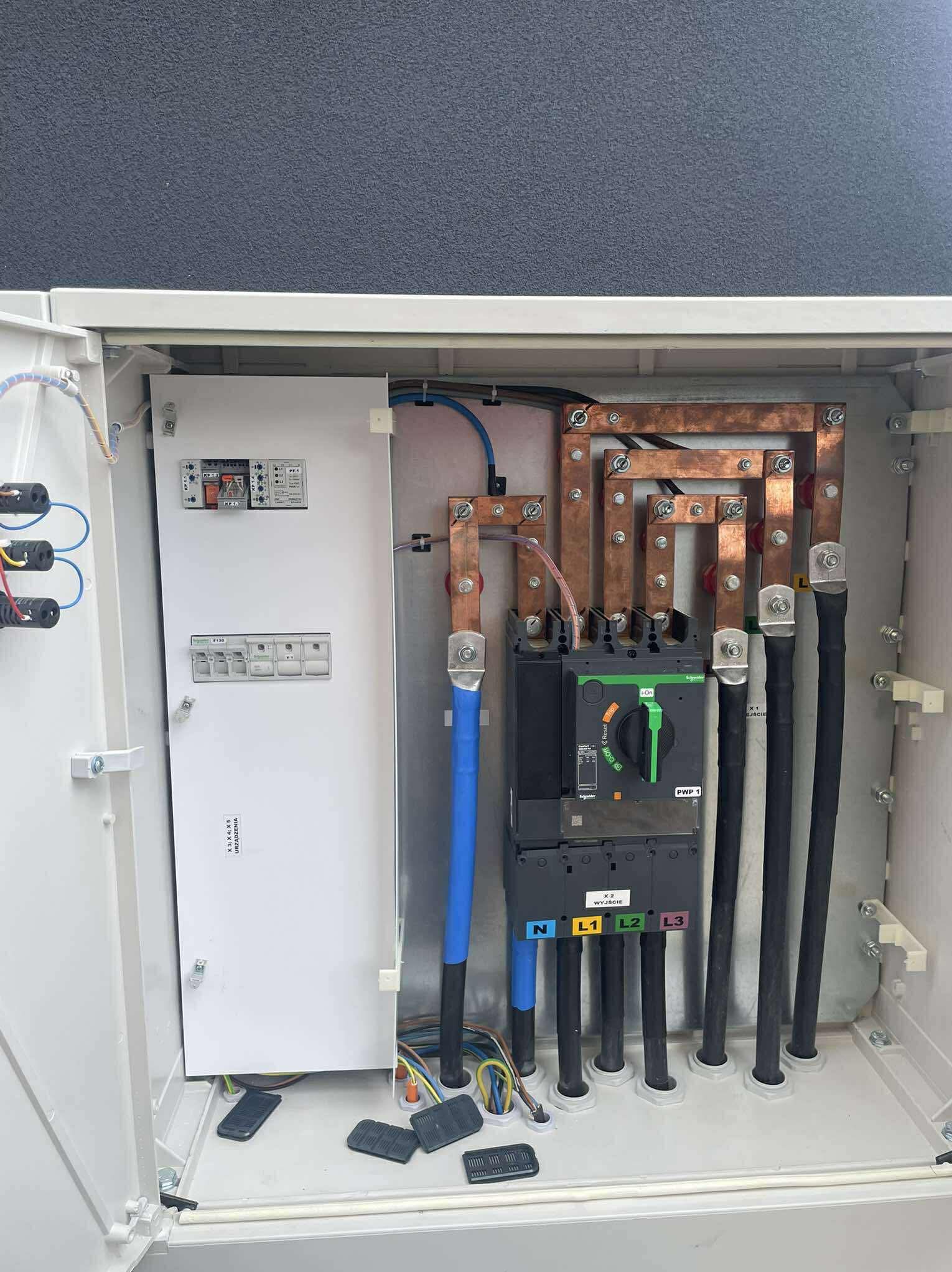 Otwarta skrzynka z zamontowaną aparaturą elektryczną: wyłączniki, szyny miedziane, przewody. Widoczne oznaczenia L1, L2, L3, N. Elementy instalacji wewnątrz obudowy.