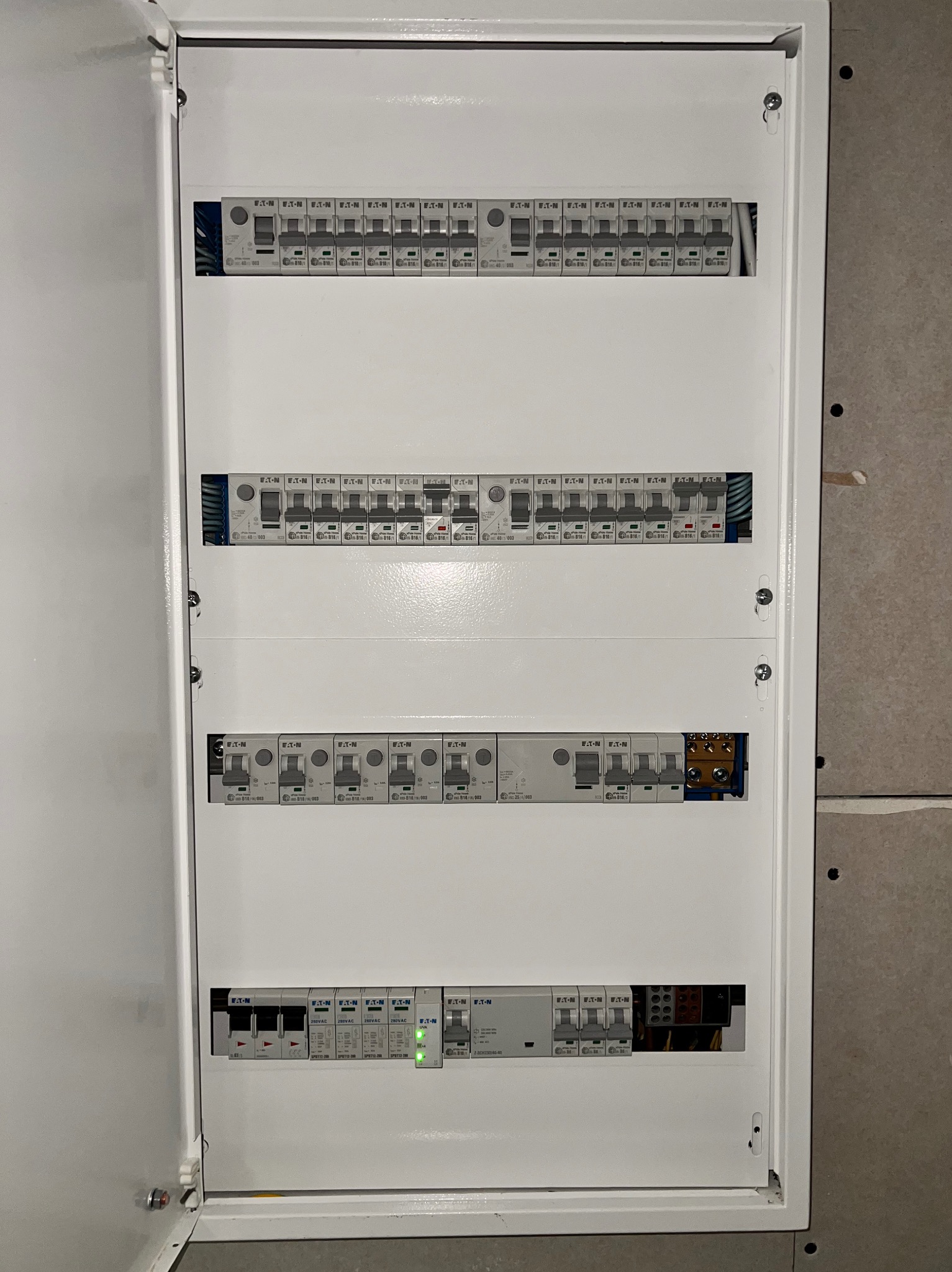 Otwarta, biała skrzynka rozdzielcza z bezpiecznikami Eaton, zamontowana na szarej ścianie. Widoczne rzędy bezpieczników i okablowanie. Jeden z bezpieczników świeci na zielono.
