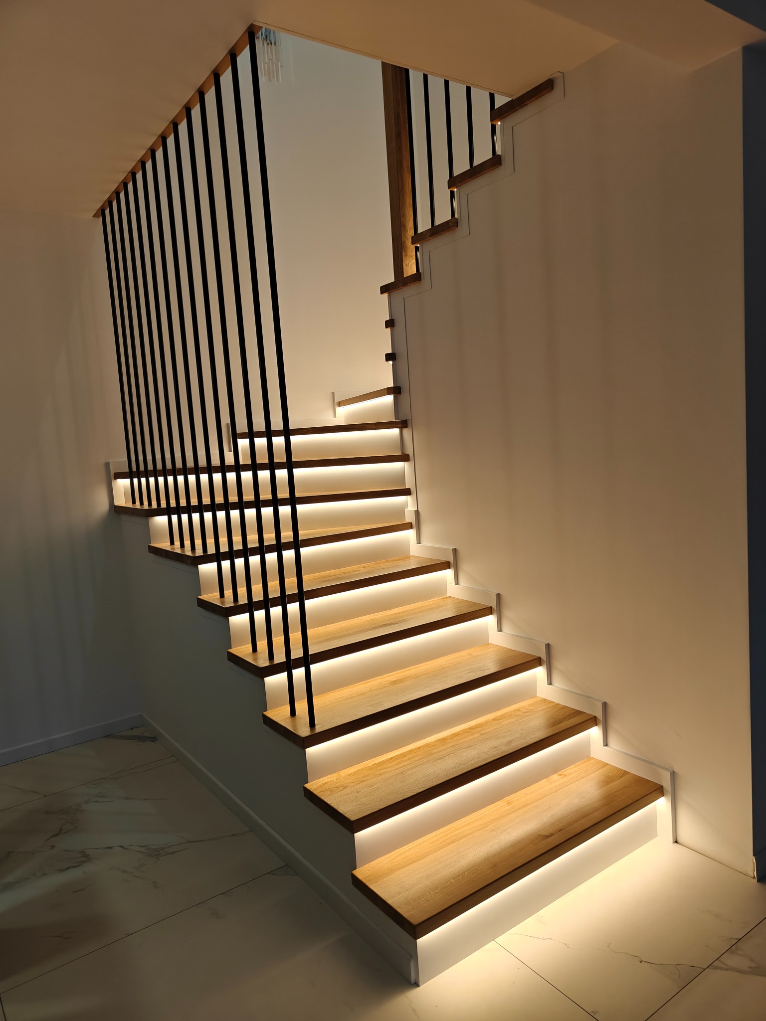 Nowoczesne schody z podświetleniem LED pod stopniami i czarną, minimalistyczną balustradą. Jasne drewno kontrastuje z bielą ścian i marmurową podłogą.