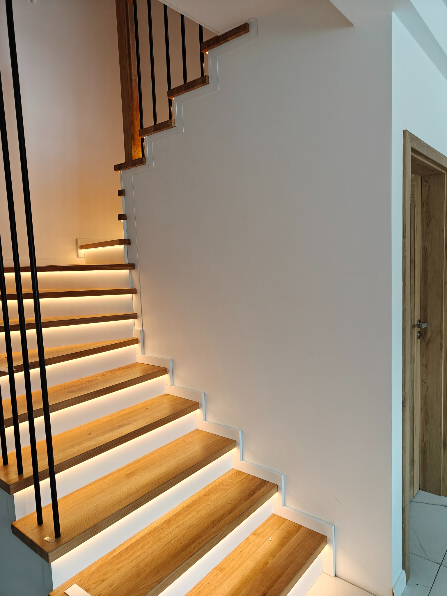 Nowoczesne schody z podświetleniem LED, drewniane stopnie i czarne metalowe balustrady. Minimalistyczny design, jasne wnętrze, widoczny fragment drewnianych drzwi.