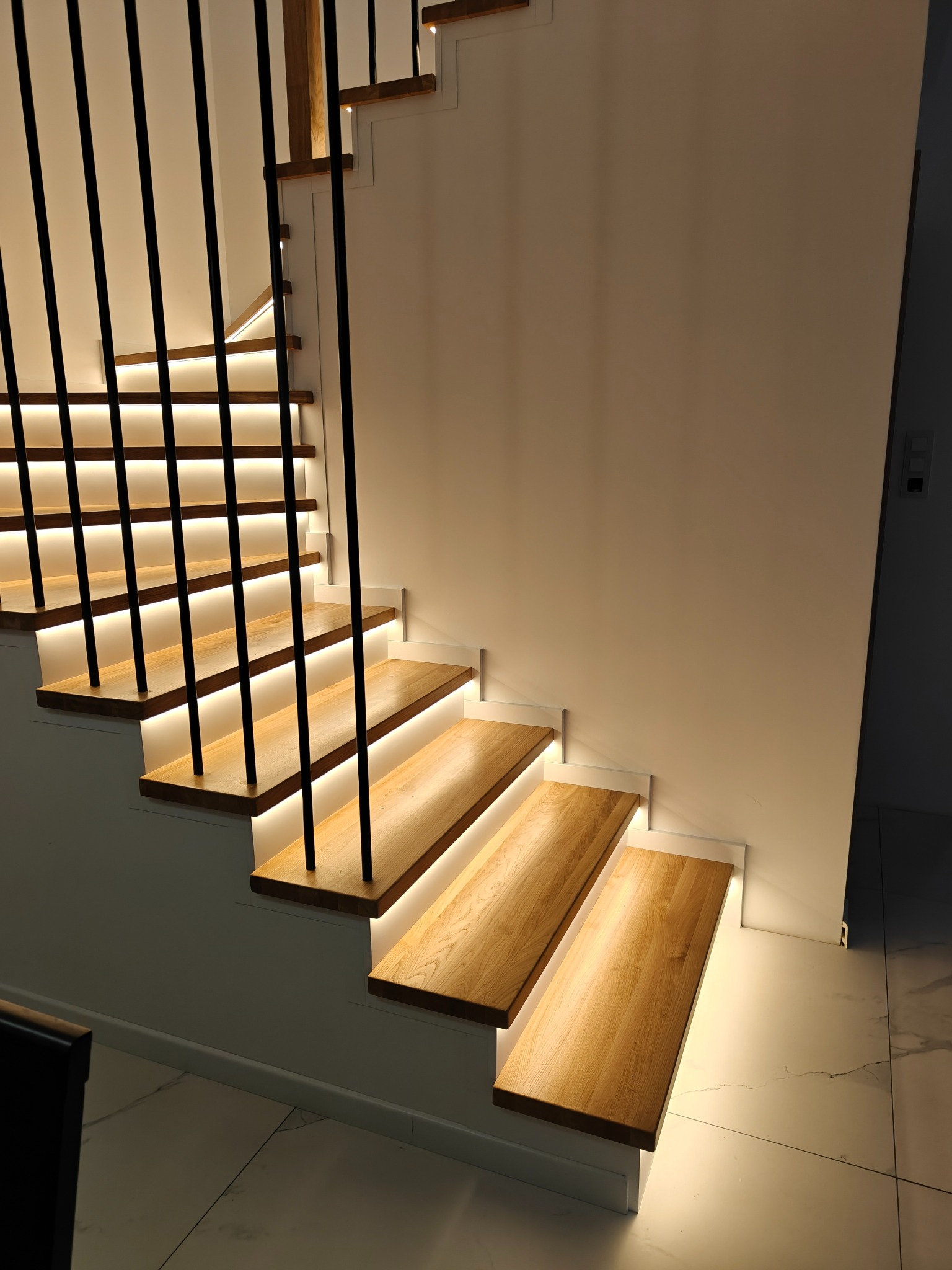 Nowoczesne schody drewniane z podświetleniem LED, czarne metalowe balustrady, jasne drewno, minimalistyczny design, białe ściany, marmurowa podłoga.
