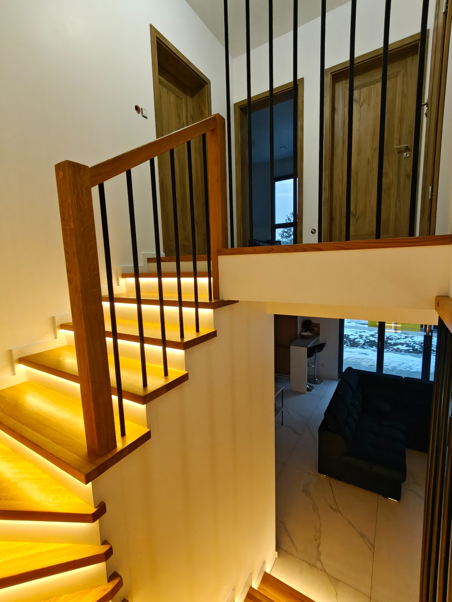 Nowoczesne schody z podświetleniem LED, drewniane stopnie i czarne metalowe balustrady. Widok z góry na salon z sofą i marmurową podłogą. Dwa drewniane drzwi w tle.