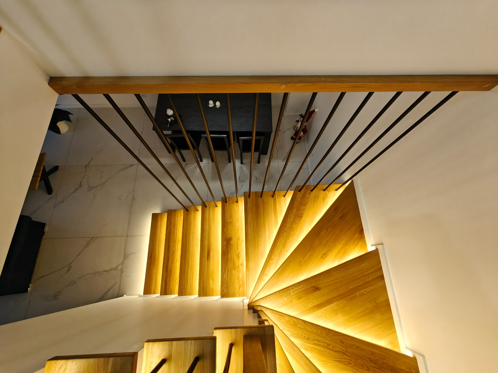 Spiralne schody z podświetlanymi stopniami, widok z góry. Metalowa balustrada z drewnianym elementem. W tle widoczny stół z krzesłami i marmurowa podłoga.
