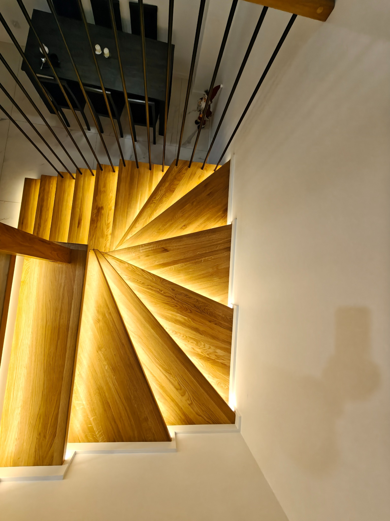 Spiralne, podświetlane stopnie z jasnego drewna, widziane z góry, z czarną balustradą i fragmentem stołu w tle. Nowoczesny design, ciepłe oświetlenie.