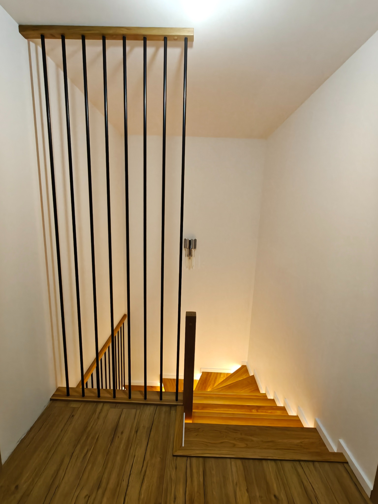 Nowoczesne schody z drewnianymi stopniami i czarną balustradą z pionowych prętów, oświetlone od dołu, widok z góry na drewnianą podłogę.