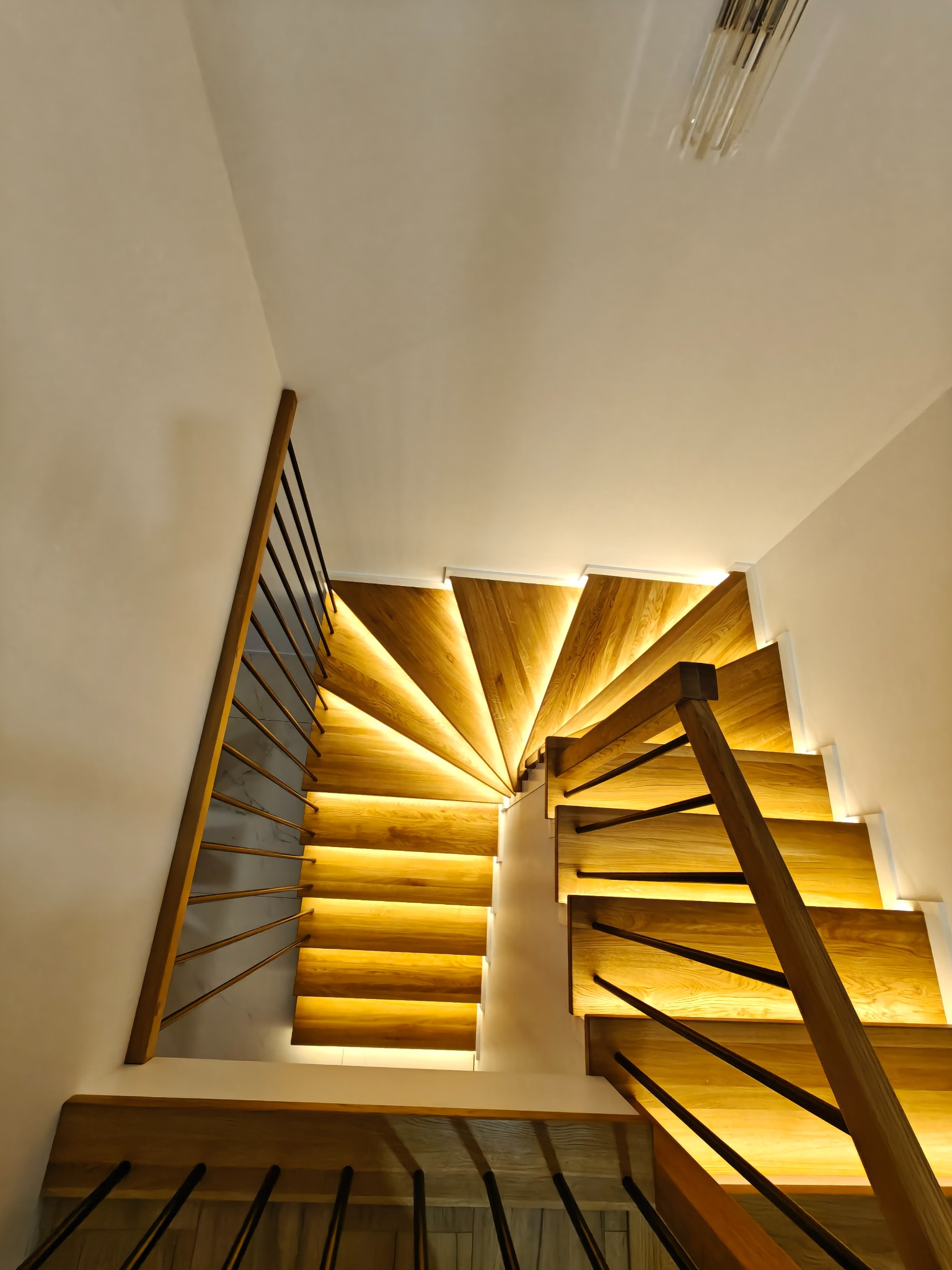 Nowoczesne schody drewniane z podświetleniem LED, widok z dołu, ukazujący spiralny układ stopni i metalową balustradę. Ciepłe światło podkreśla strukturę drewna.