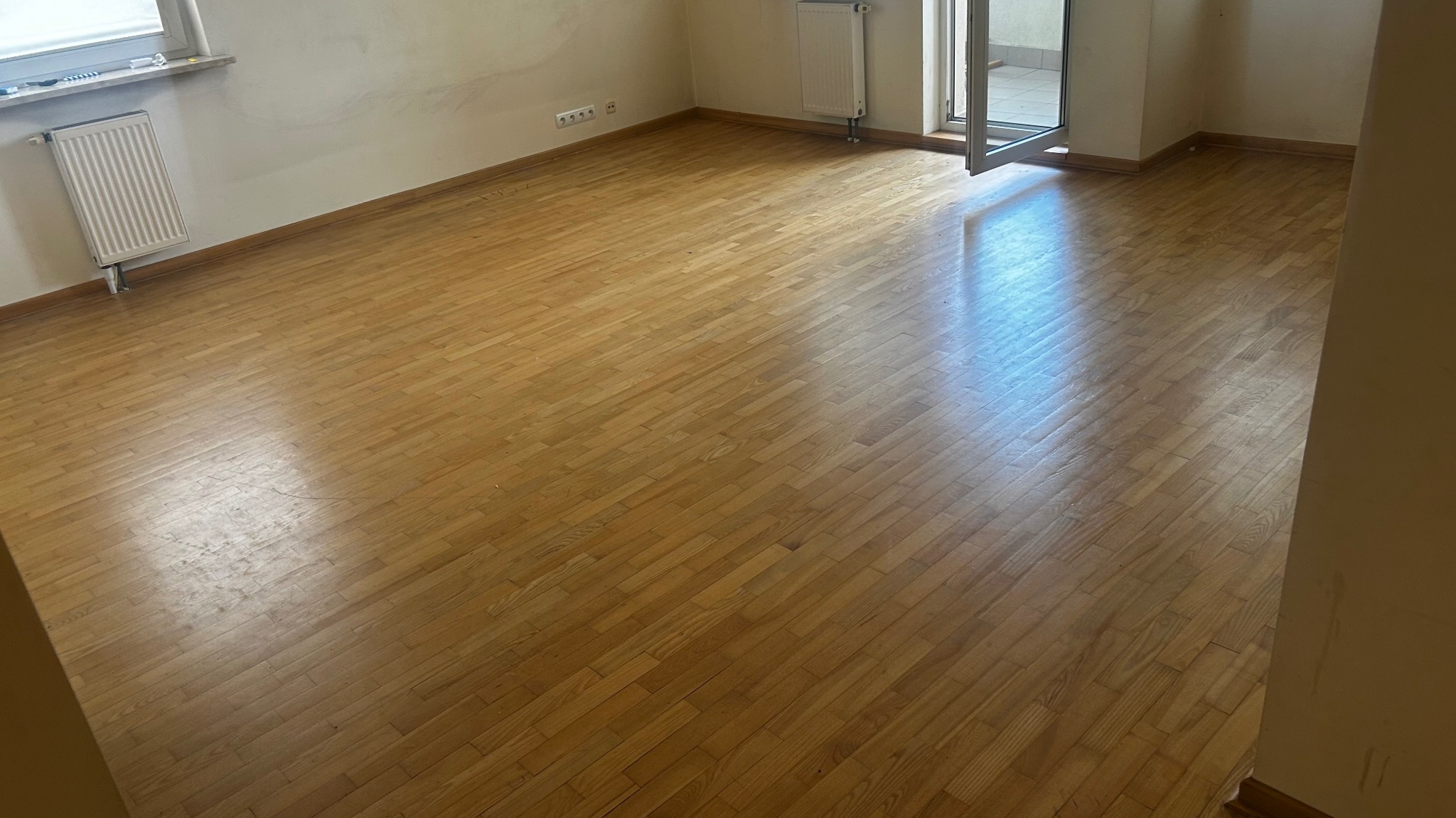 Jasna, odnowiona podłoga parkietowa w pustym pokoju z grzejnikami i otwartym balkonem. Widoczne ślady po remoncie na ścianach.