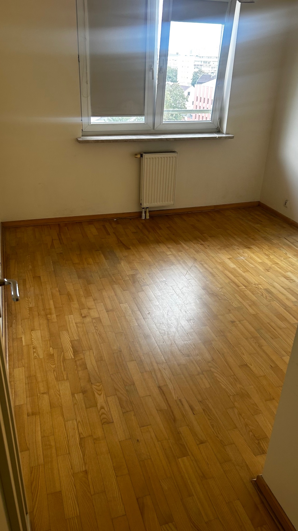 Pokój z parkietem w jodełkę, okno z roletą i grzejnik. Widok na budynki za oknem. Podłoga po renowacji, gotowa do dalszych prac wykończeniowych.