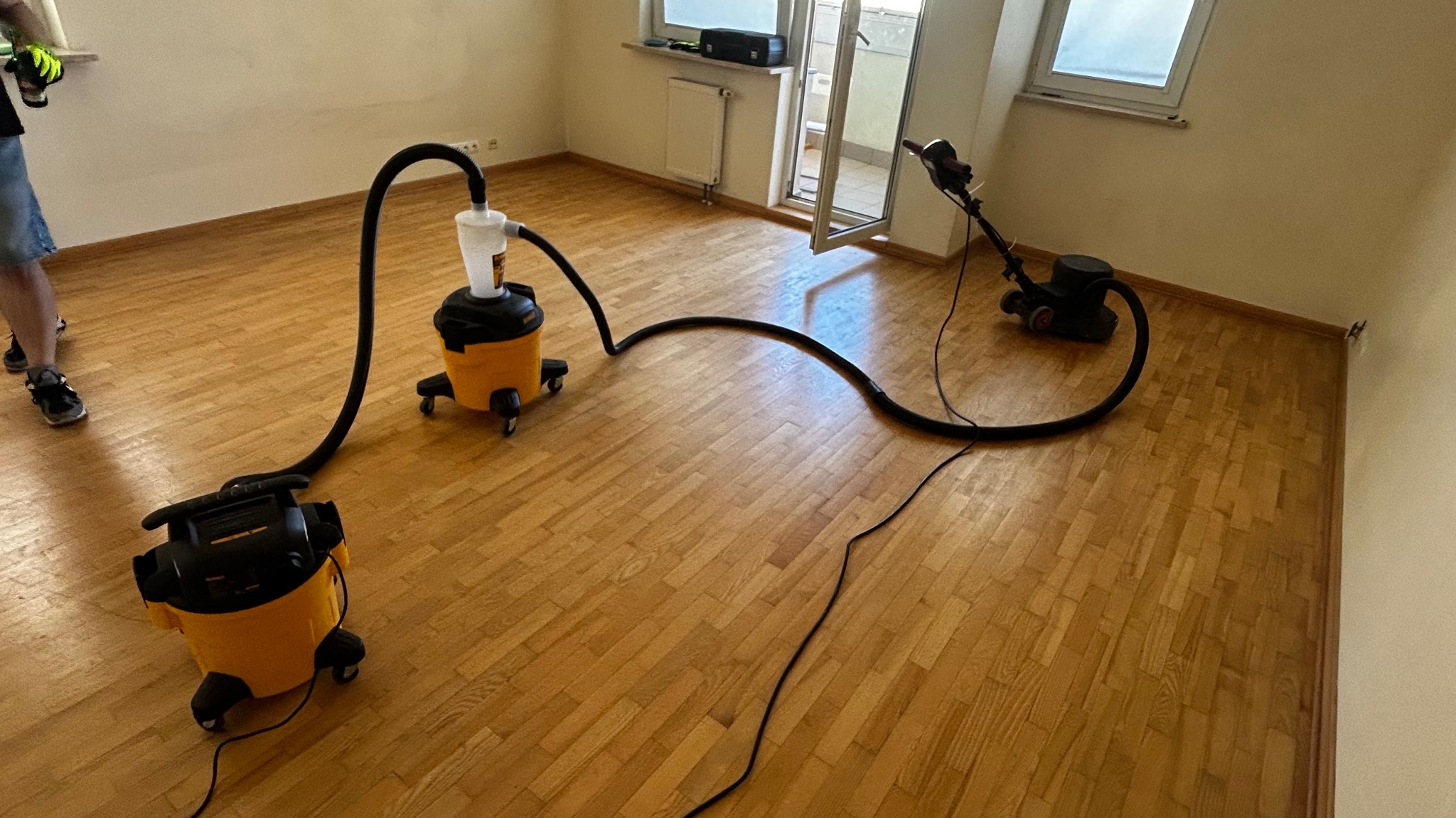 Parkiet po cyklinowaniu z widocznymi maszynami do obróbki drewna i odkurzaczami przemysłowymi. Jasne drewno, naturalne światło, widok na balkon.