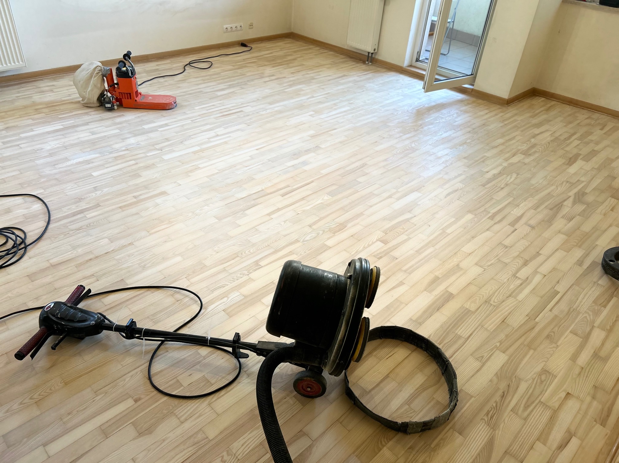 Jasna, odnowiona podłoga parkietowa w pokoju z cykliniarką i szlifierką. Widoczne ślady po cyklinowaniu. Otwarty balkon w tle.