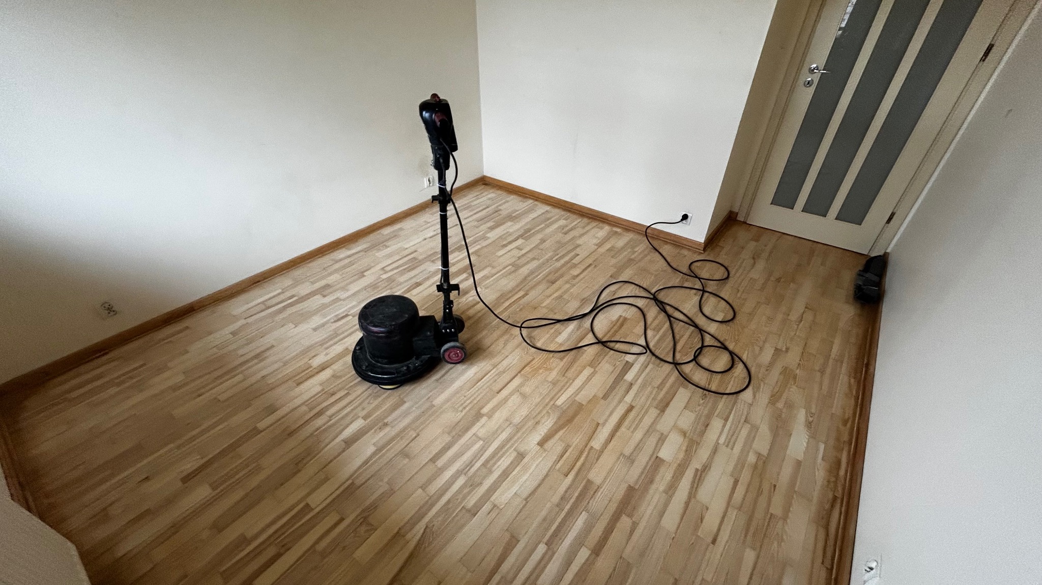 Cyklinowanie parkietu w rogu pokoju. Widoczna maszyna do cyklinowania z długim kablem. Świeżo wycyklinowana podłoga z jasnego drewna.