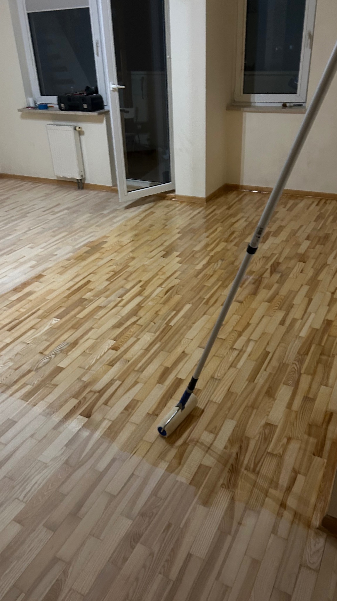 Lakierowanie parkietu wałkiem w jasnym pomieszczeniu. Część podłogi już polakierowana, widoczna różnica w odcieniu drewna. Okno i drzwi balkonowe w tle.