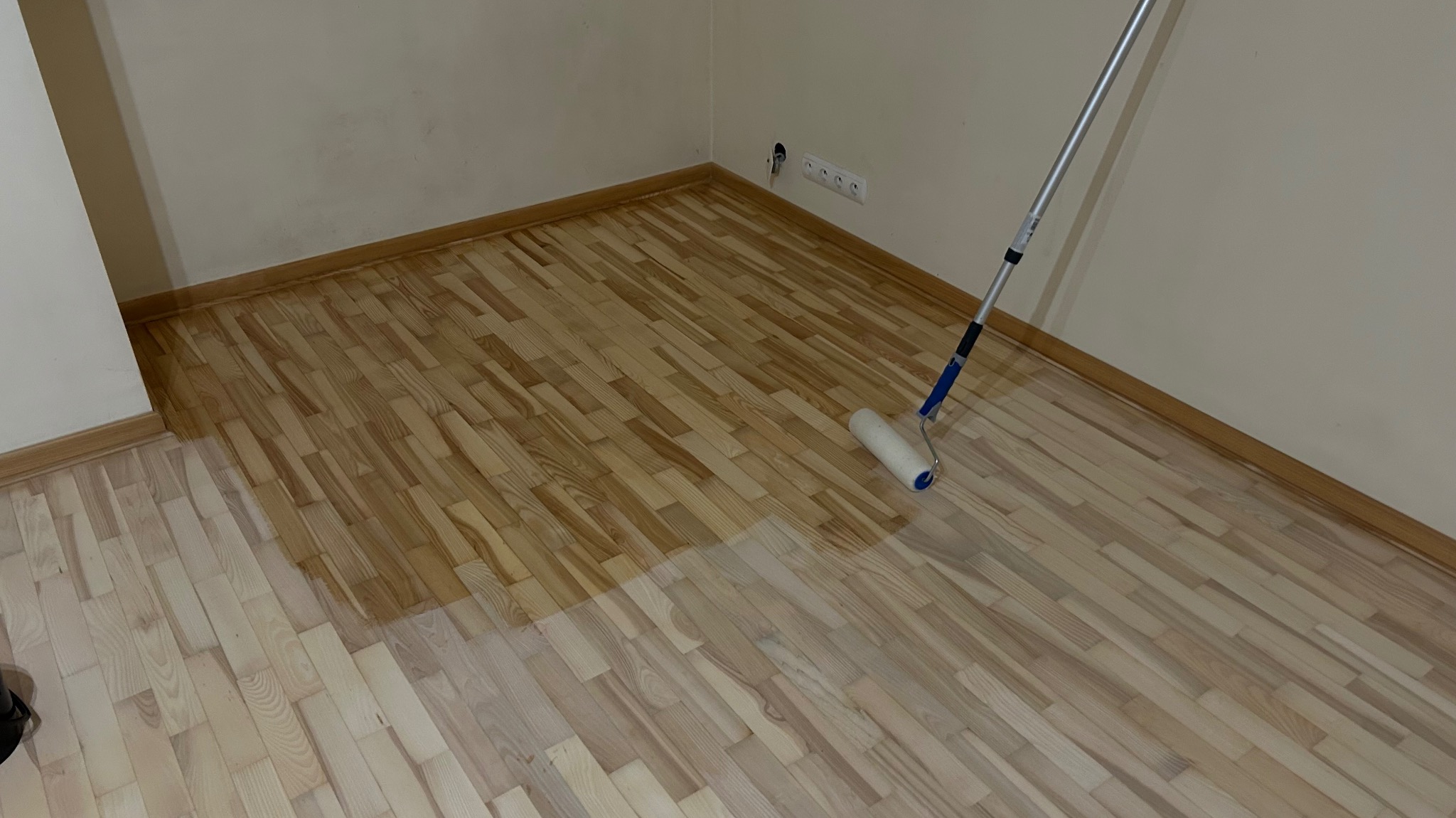 Parkiet po cyklinowaniu i lakierowaniu. Część podłogi pokryta lakierem, widoczny wałek malarski. Jasne drewno, gładka powierzchnia, świeżo po renowacji.