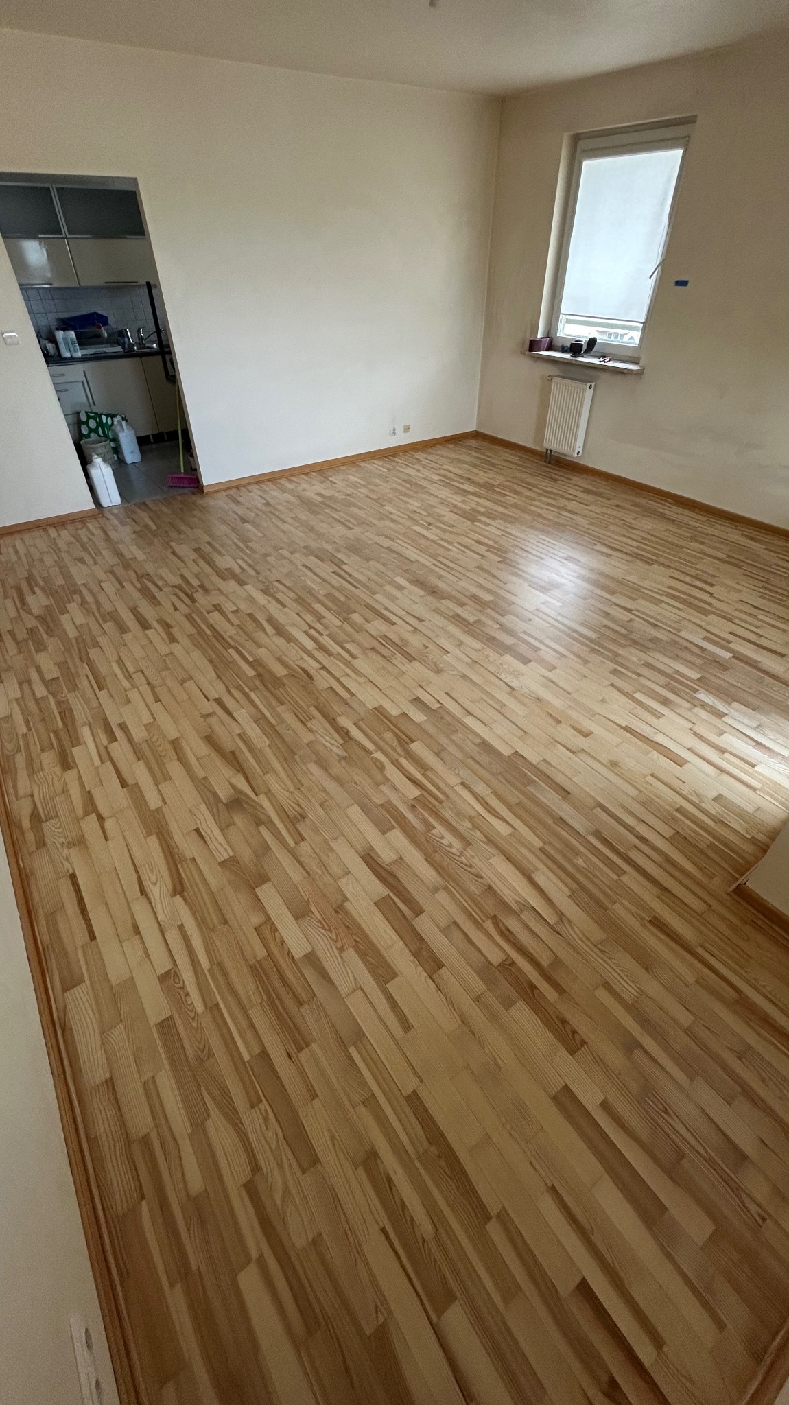 Jasna, odnowiona drewniana podłoga parkietowa w pustym pokoju z oknem i widokiem na kuchnię. Świeżo wycyklinowany parkiet w naturalnym odcieniu drewna.