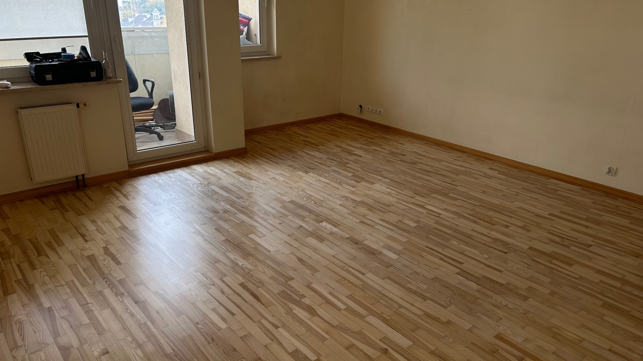 Jasna, odnowiona podłoga parkietowa w pustym pokoju z balkonem. Widoczne listwy przypodłogowe i gniazdka elektryczne. Na balkonie krzesło biurowe.