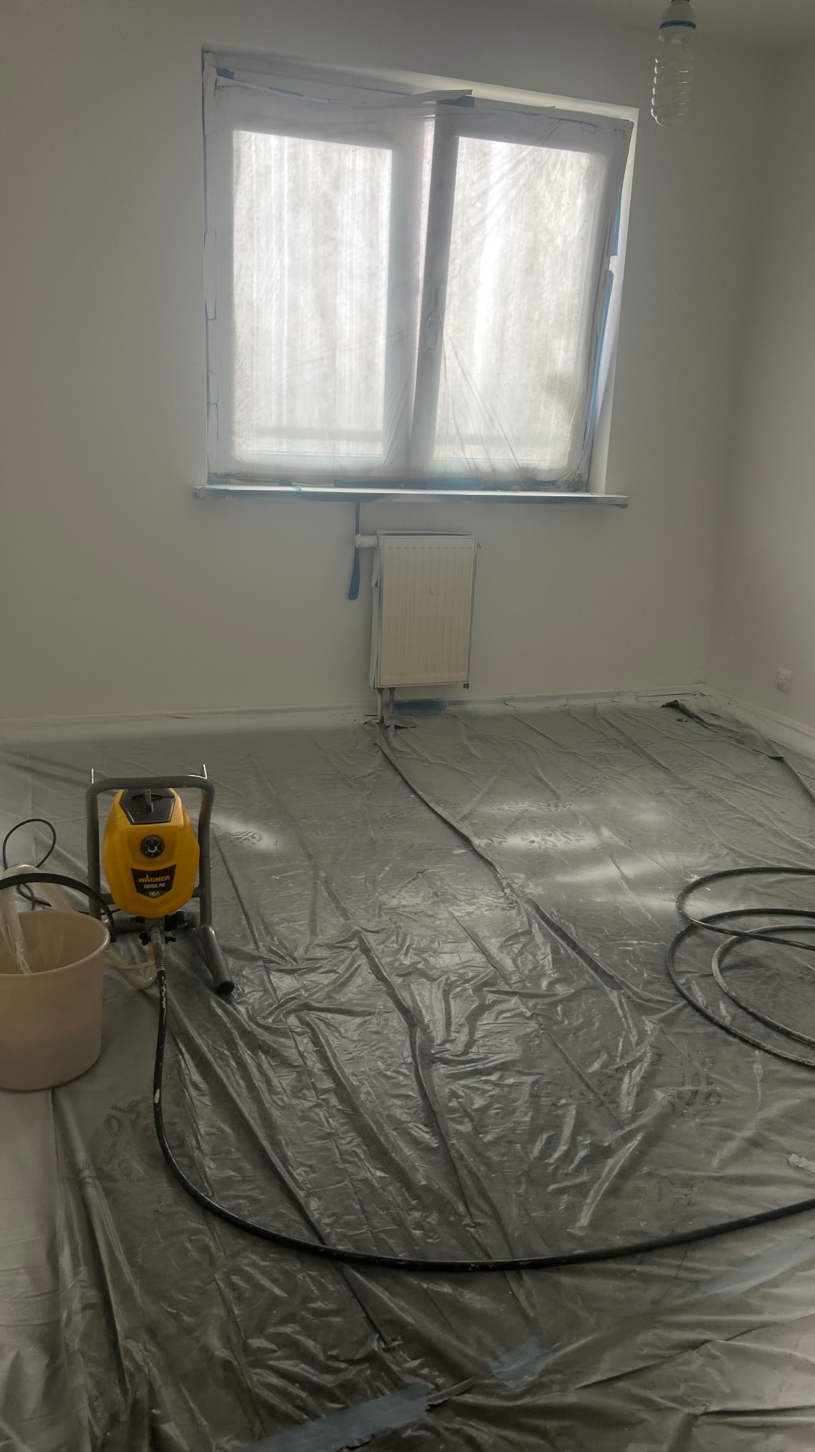 Pokój przygotowany do malowania natryskowego: okno i grzejnik zabezpieczone folią, podłoga przykryta folią ochronną, widoczny agregat malarski.