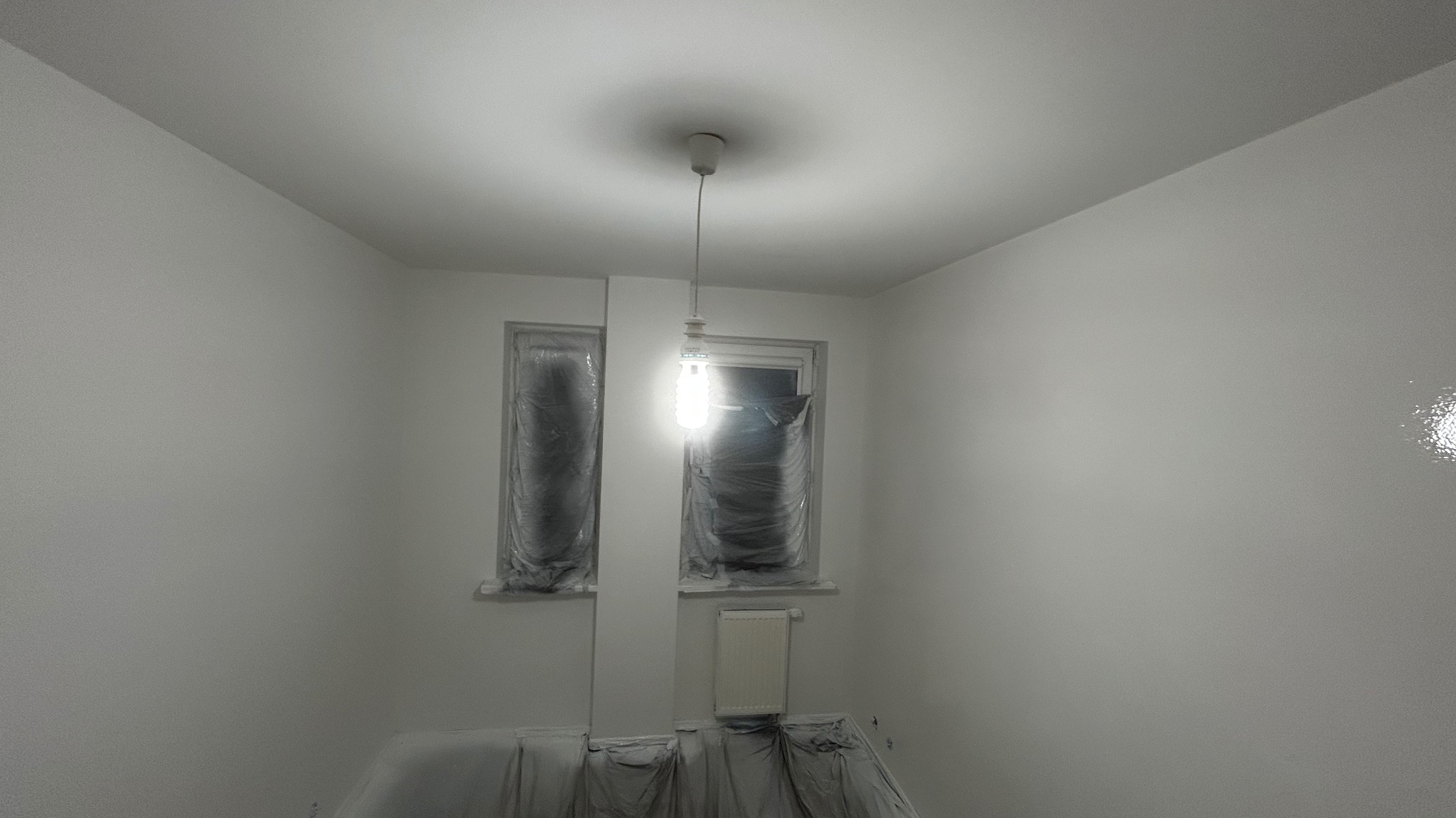 Białe, świeżo pomalowane wnętrze w Warszawie. Okna i podłoga zabezpieczone folią malarską. Widoczna żarówka. Świeżo po malowaniu.
