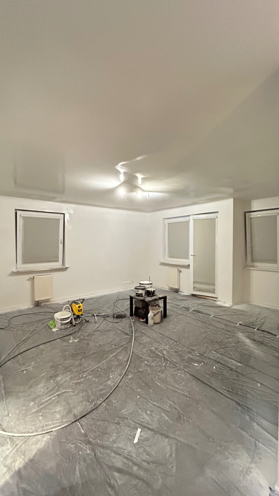 Przygotowanie do malowania: Pokój zabezpieczony folią, widoczny agregat malarski, farby i narzędzia na stoliku. Świeżo pomalowane ściany na biało.