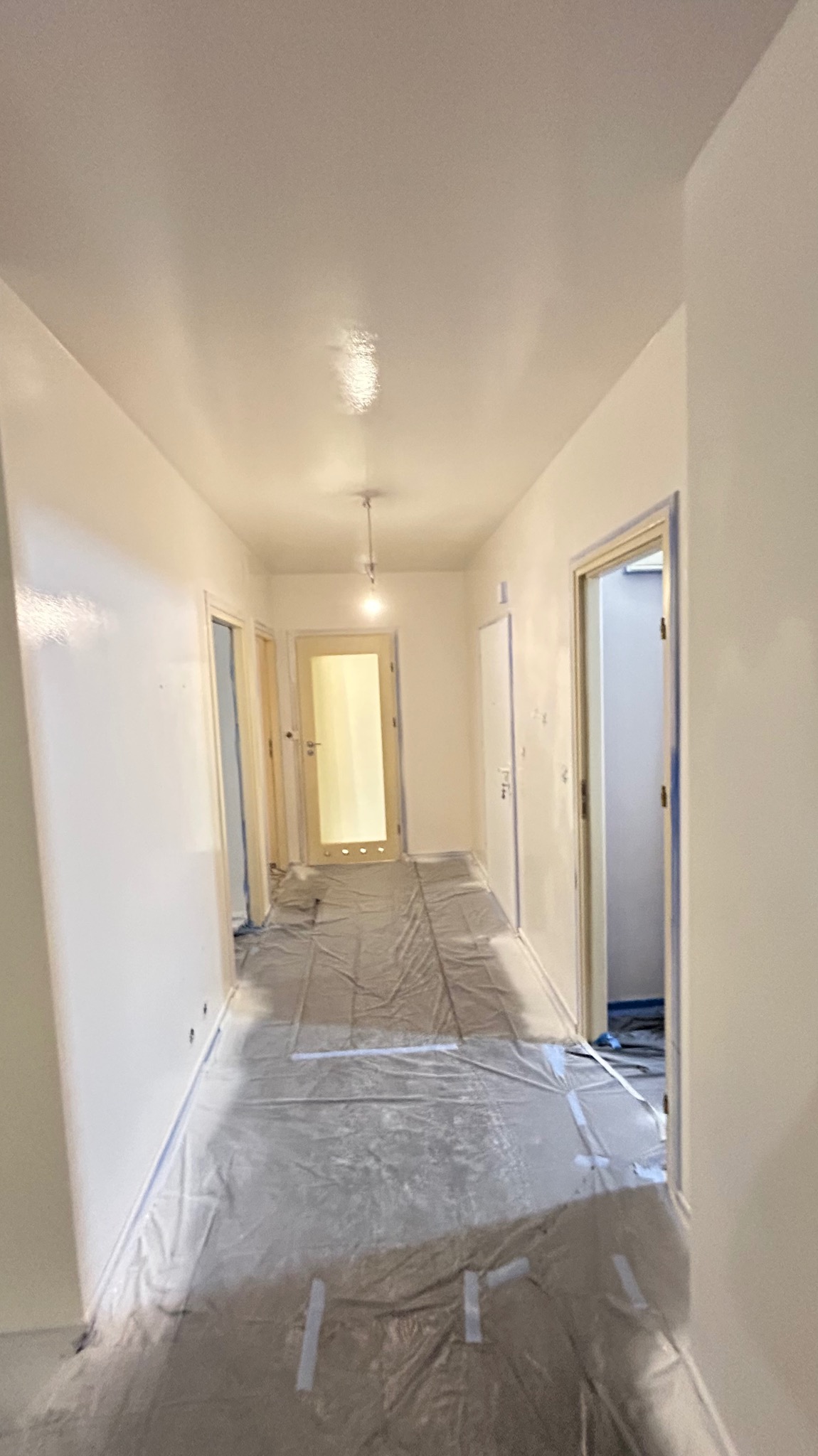 Biały korytarz w trakcie malowania, zabezpieczony folią malarską. Widoczne świeżo pomalowane ściany i sufit, taśmy malarskie przy futrynach drzwi.