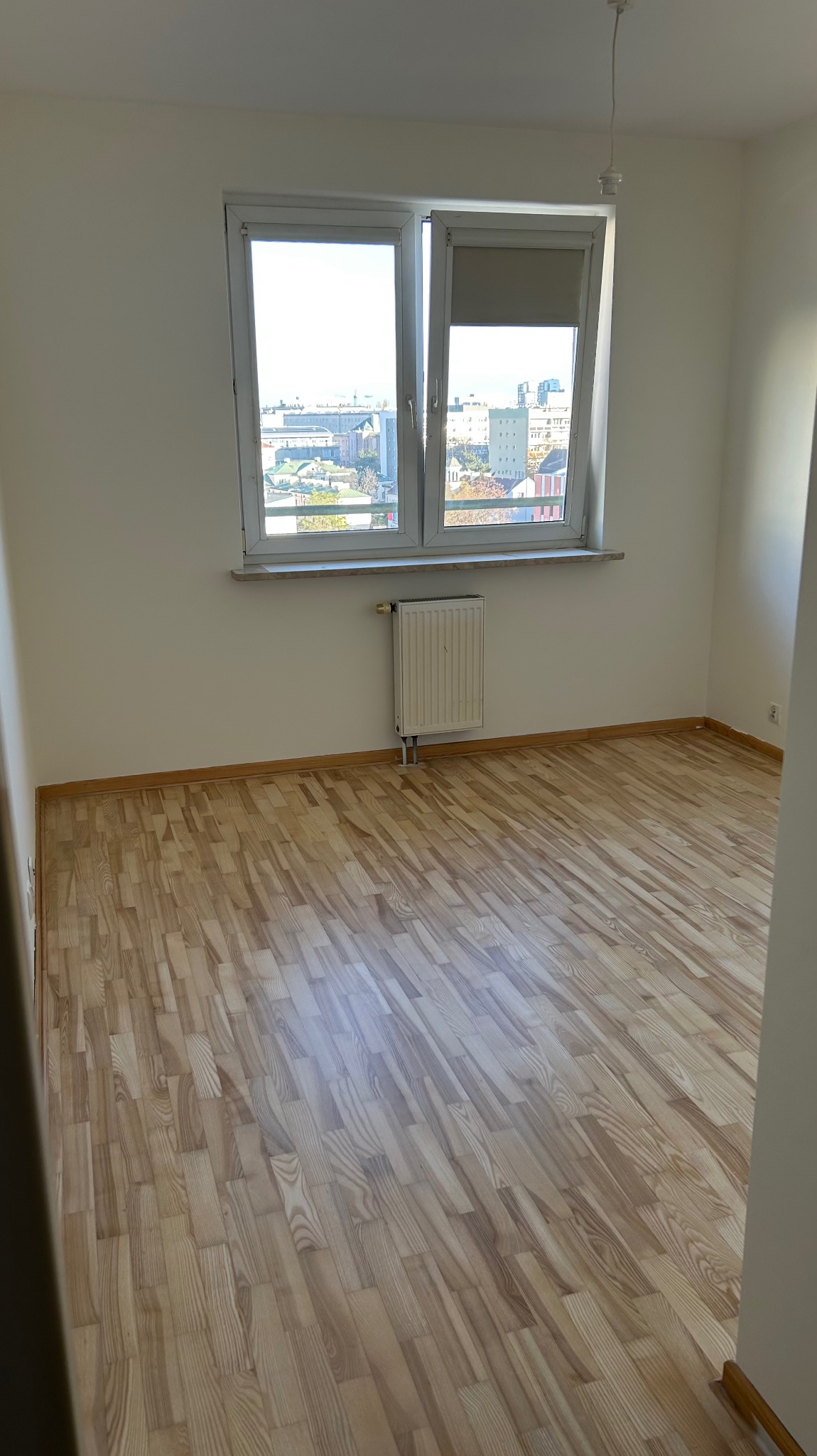 Jasne, parkietowe panele podłogowe w pustym pokoju z oknem i grzejnikiem. Widok na miasto za oknem. Świeżo wykończone wnętrze, gotowe do umeblowania.