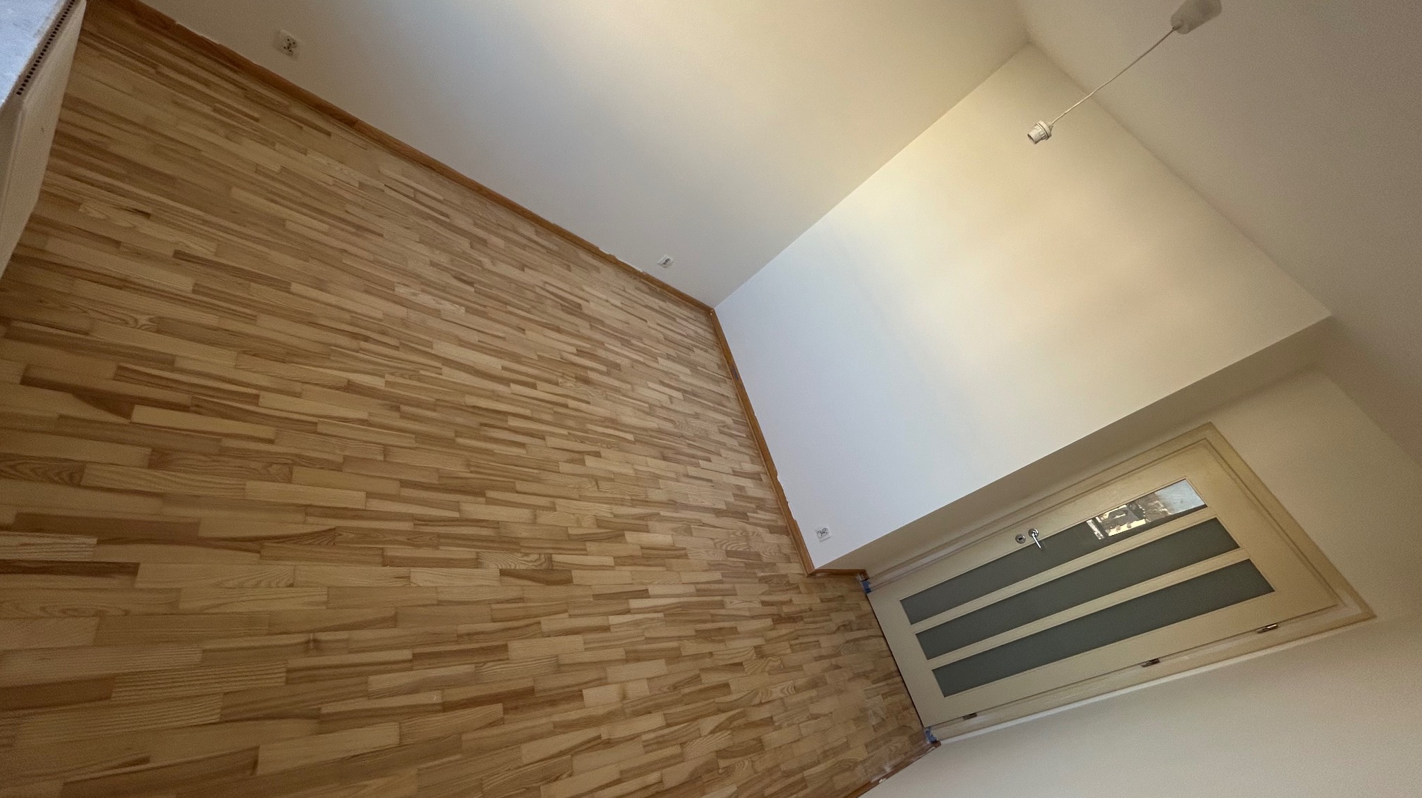 Jasna, drewniana podłoga parkietowa ułożona w jodełkę, świeżo po renowacji, widoczna z perspektywy pod kątem, z fragmentem białej ściany i drzwi w tle.