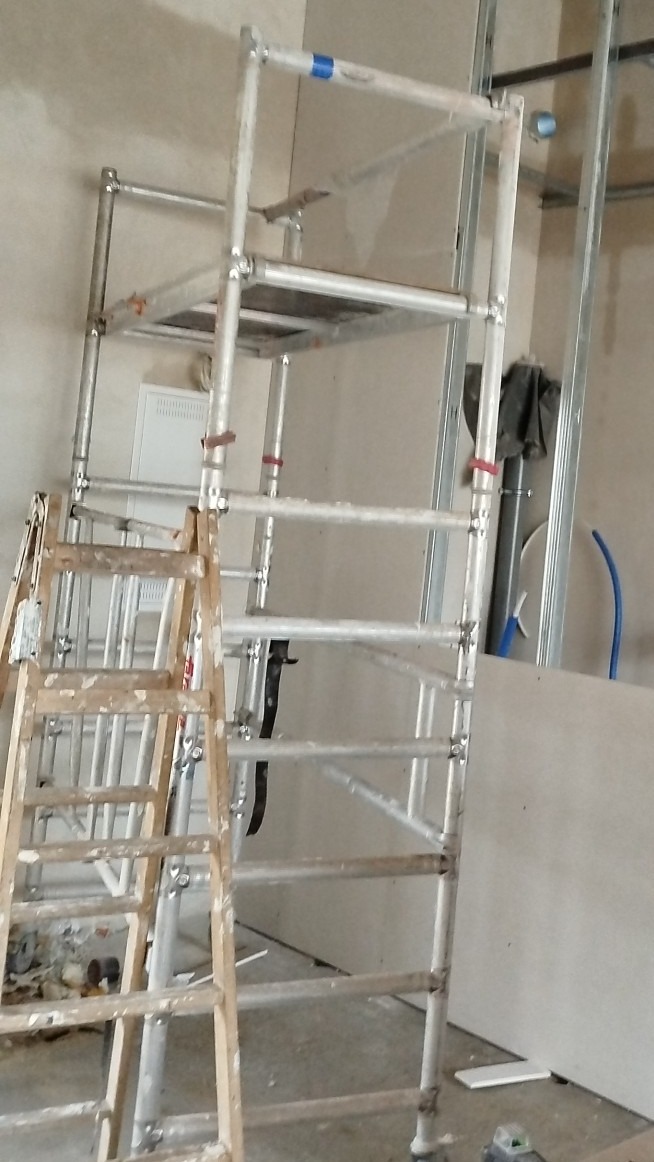 Wnętrze w trakcie prac wykończeniowych: aluminiowe rusztowanie z drewnianą drabiną, widoczne profile do płyt gipsowo-kartonowych i instalacje.