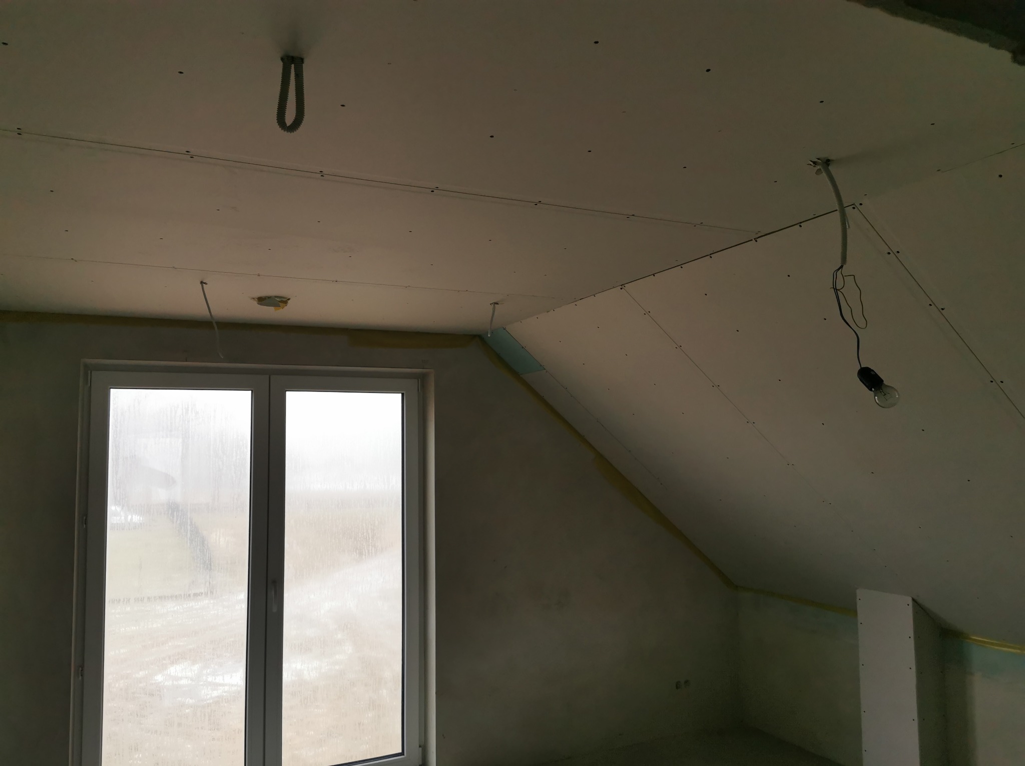 Poddasze w trakcie wykańczania płytami karton-gips, widoczne łączenia i okno z widokiem na zewnątrz. W tle surowe ściany i prowizoryczne oświetlenie.