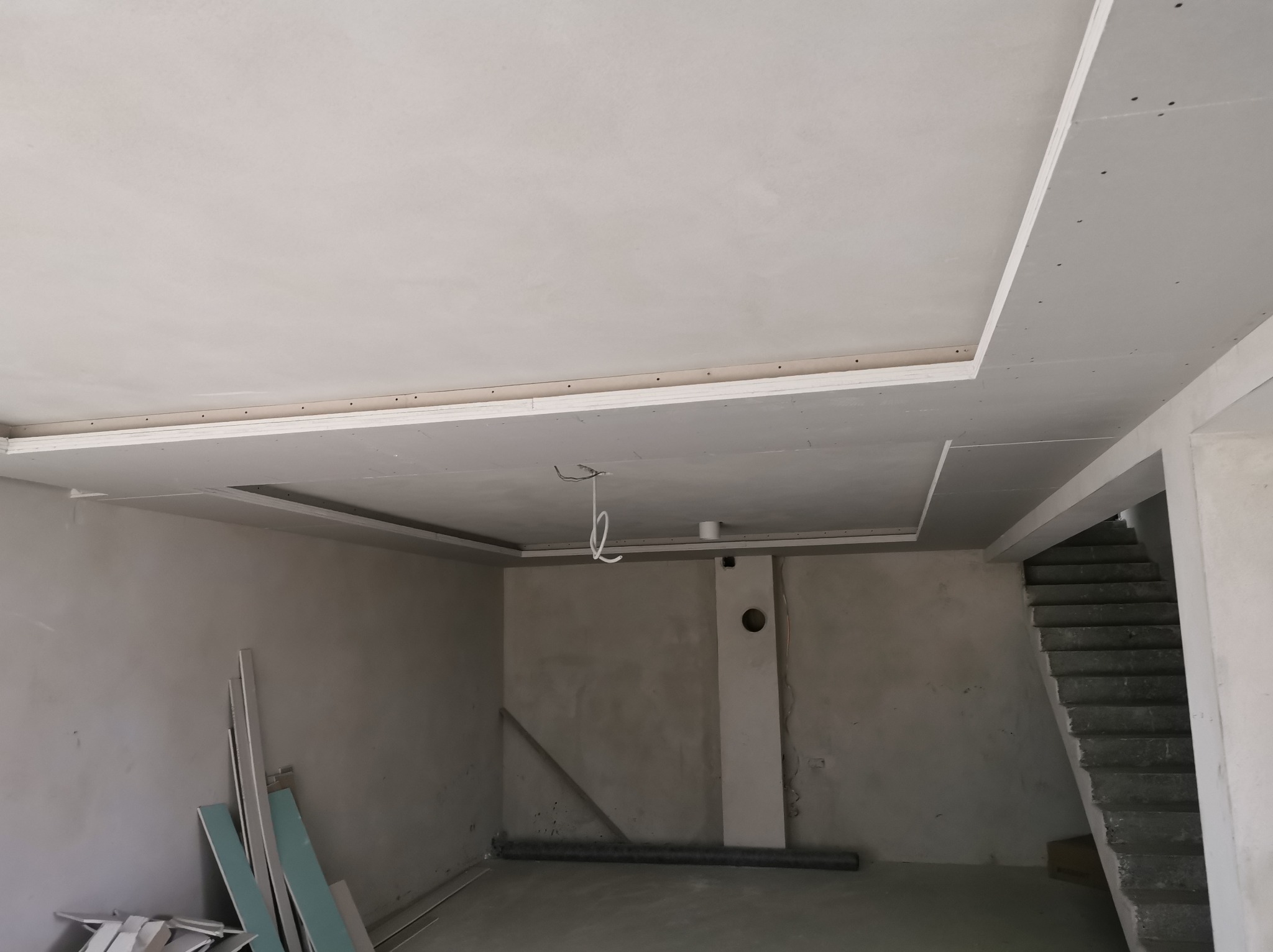 Wykończony sufit z płyt kartonowo-gipsowych z ozdobnym obniżeniem w nowym domu. Widoczne płyty gipsowe i przewody elektryczne. Schody w tle.