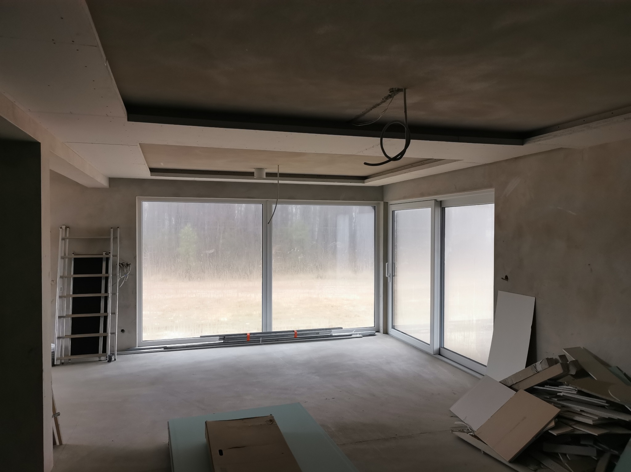 Wnętrze w trakcie wykończenia: surowy sufit z płyt karton-gips, duże okna przesuwne, drabina i materiały budowlane. Widoczne kable i niedokończone prace.