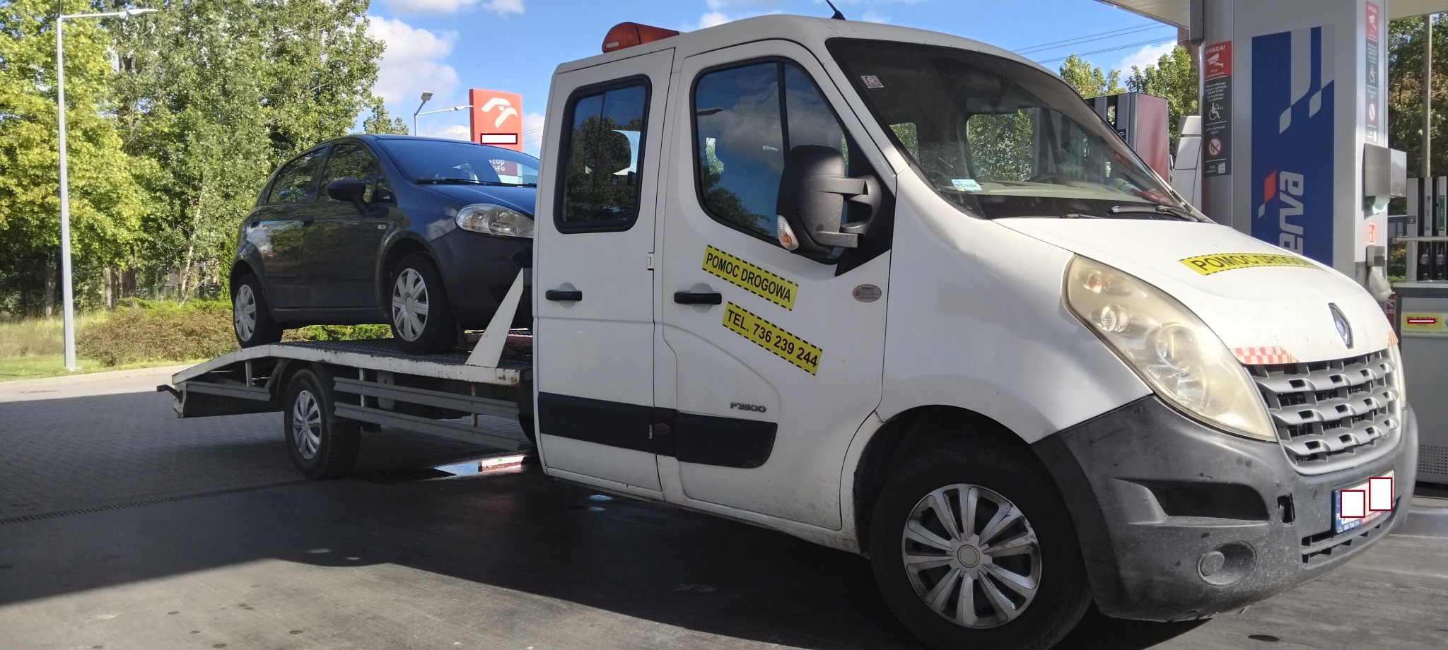 Biała laweta Renault z czarnym autem osobowym na platformie, zaparkowana przy stacji benzynowej w słoneczny dzień. Na drzwiach naklejka 'Pomoc Drogowa'.