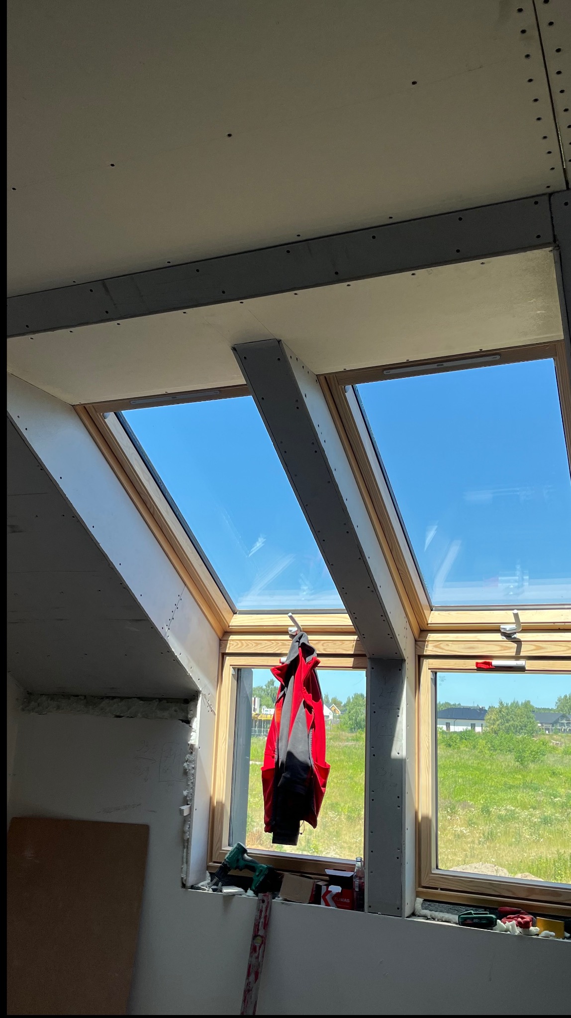 Montaż płyt gipsowo-kartonowych na poddaszu z oknami dachowymi. Widoczne profile, płyty, narzędzia i częściowo widoczny krajobraz za oknem. Prace wykończeniowe w jasnym wnętrzu.