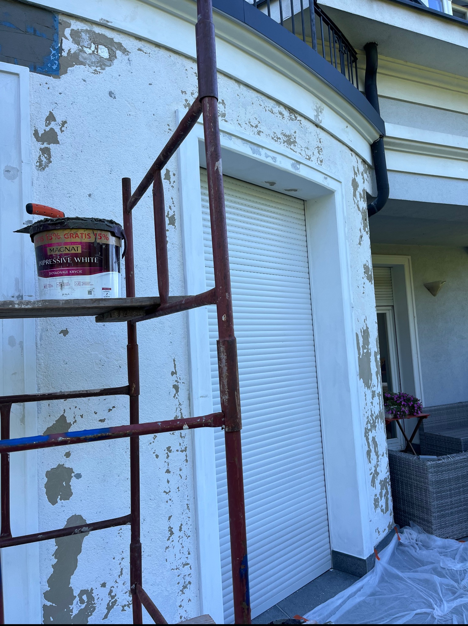 Renowacja elewacji: rusztowanie z farbą Magnat Impressive White przy ścianie z odpryskami, przygotowanej do malowania. Widoczne zabezpieczenia.