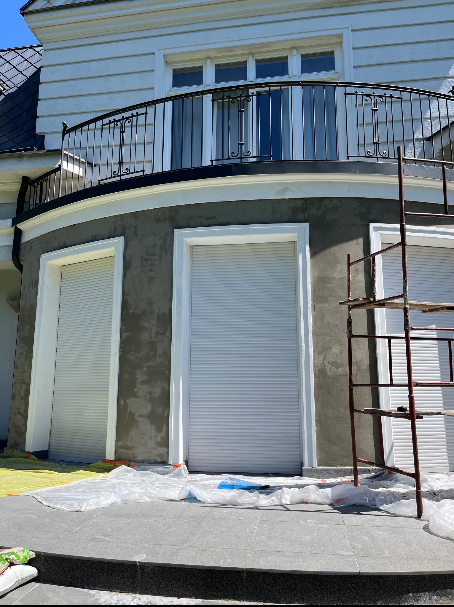 Elewacja budynku z oknami z roletami zewnętrznymi w trakcie prac wykończeniowych. Widoczne rusztowanie i folie ochronne. Świeżo nałożona warstwa szpachli.