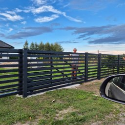 Tomasz Śliwiński MEXA-STYL - Nowoczesne, poziome ogrodzenie metalowe w kolorze antracytowym, z widocznymi dwoma mężczyznami w tle, na tle domu i błękitnego nieba z chmurami.