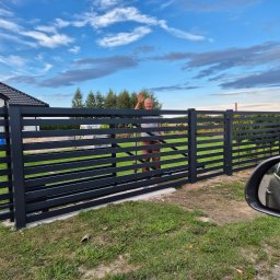 Tomasz Śliwiński MEXA-STYL - Nowoczesne, poziome ogrodzenie panelowe w kolorze grafitowym, z widoczną bramą i uśmiechniętym mężczyzną machającym ręką, na tle domu i błękitnego nieba.
