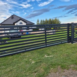 Tomasz Śliwiński MEXA-STYL - Nowoczesne, poziome ogrodzenie posesji z szarych, metalowych paneli na tle domu z ciemnym dachem i błękitnego nieba z chmurami. Widoczny mechanizm bramy przesuwnej.