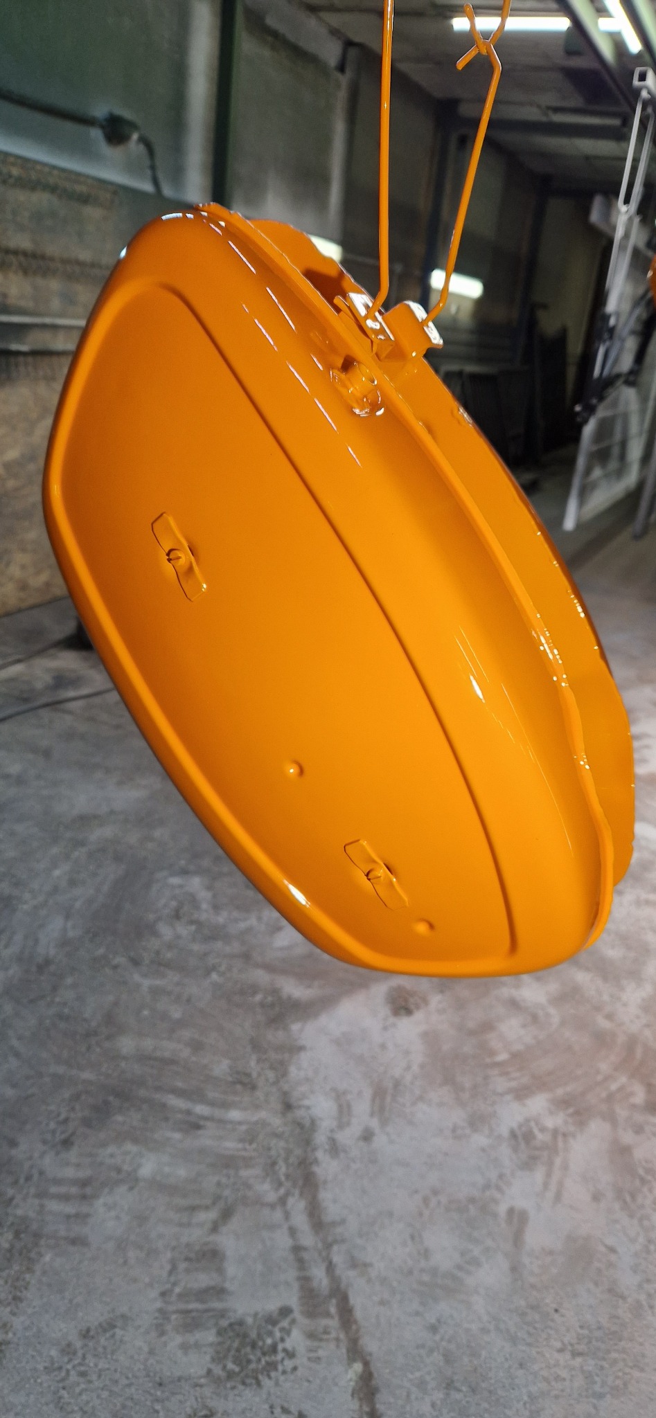 Pomarańczowy element metalowy, prawdopodobnie zbiornik, zawieszony na haku po malowaniu proszkowym. Błyszcząca powierzchnia, widoczne detale mocowań.