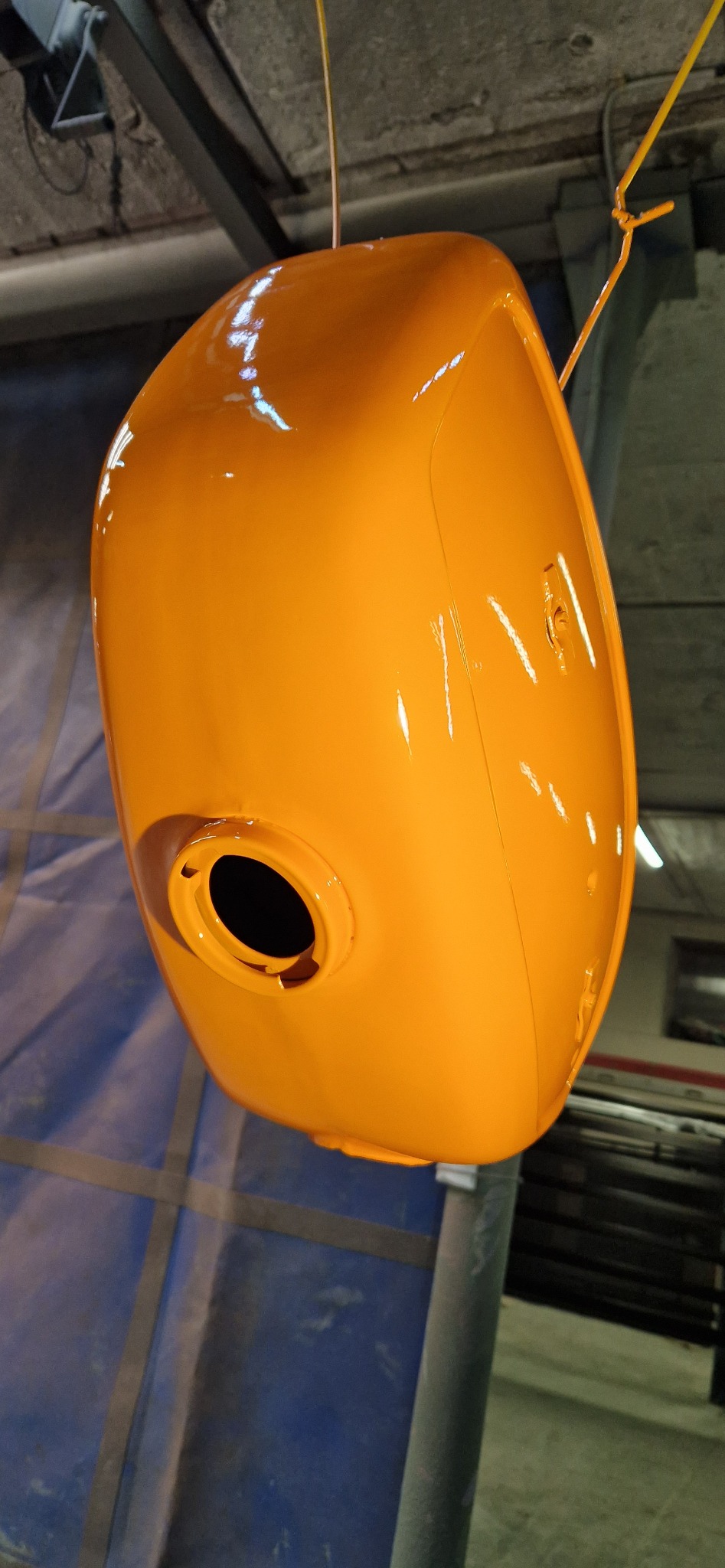 Pomarańczowy zbiornik po malowaniu proszkowym, zawieszony w warsztacie. Widoczne refleksy światła na gładkiej powierzchni. Detal z otworem w dolnej części zbiornika.