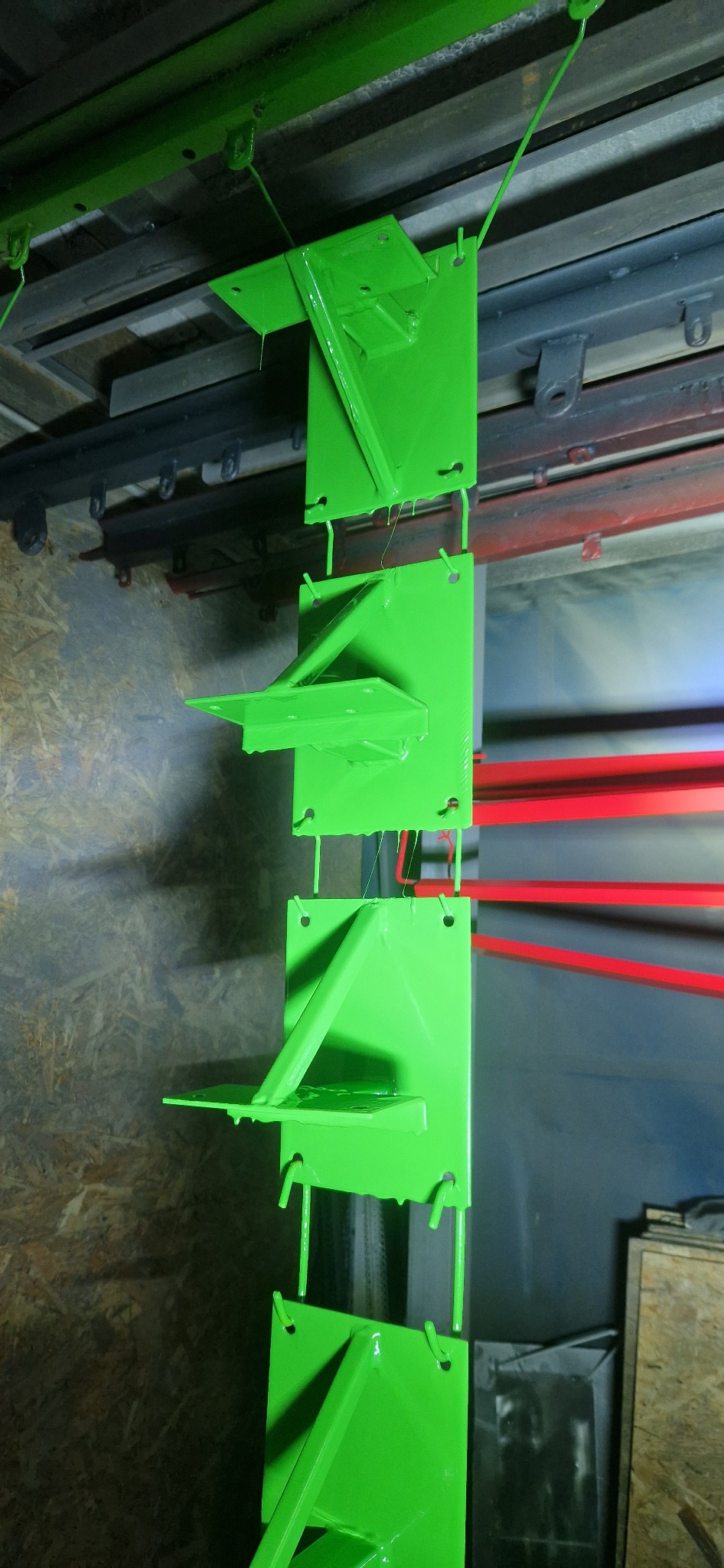 Świeżo pomalowane na jaskrawy zielony kolor metalowe elementy konstrukcyjne, wiszące na lince w hali produkcyjnej, z widocznymi śladami farby.