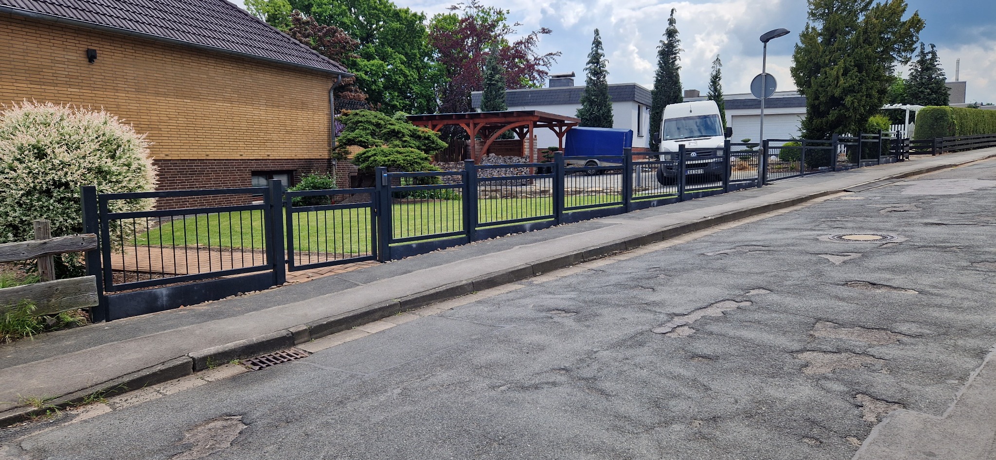 Ciemne, metalowe ogrodzenie posesji z furtką i bramą, widoczne z ulicy. W tle dom, zieleń i zaparkowany bus. Ujęcie z poziomu ulicy.