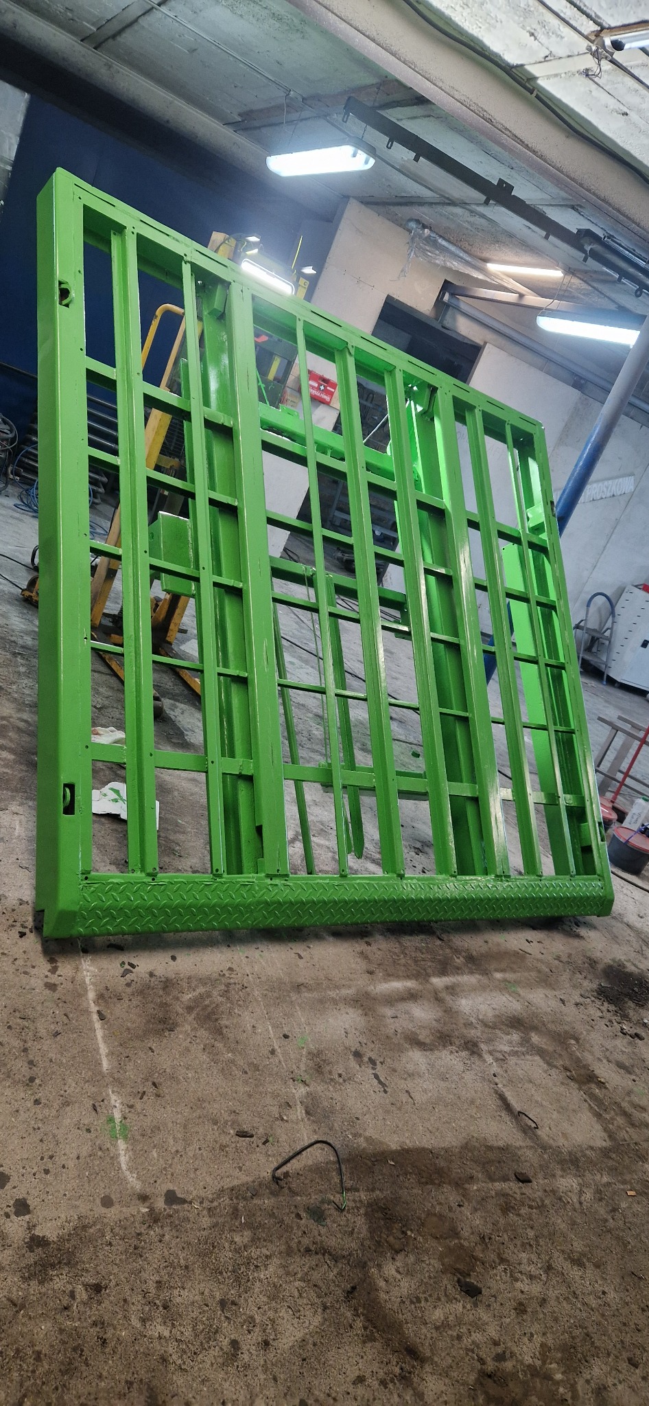 Świeżo pomalowany na zielono metalowy element konstrukcyjny z kratownicą, leżący na betonowej podłodze w hali. Widoczne ślady po malowaniu.
