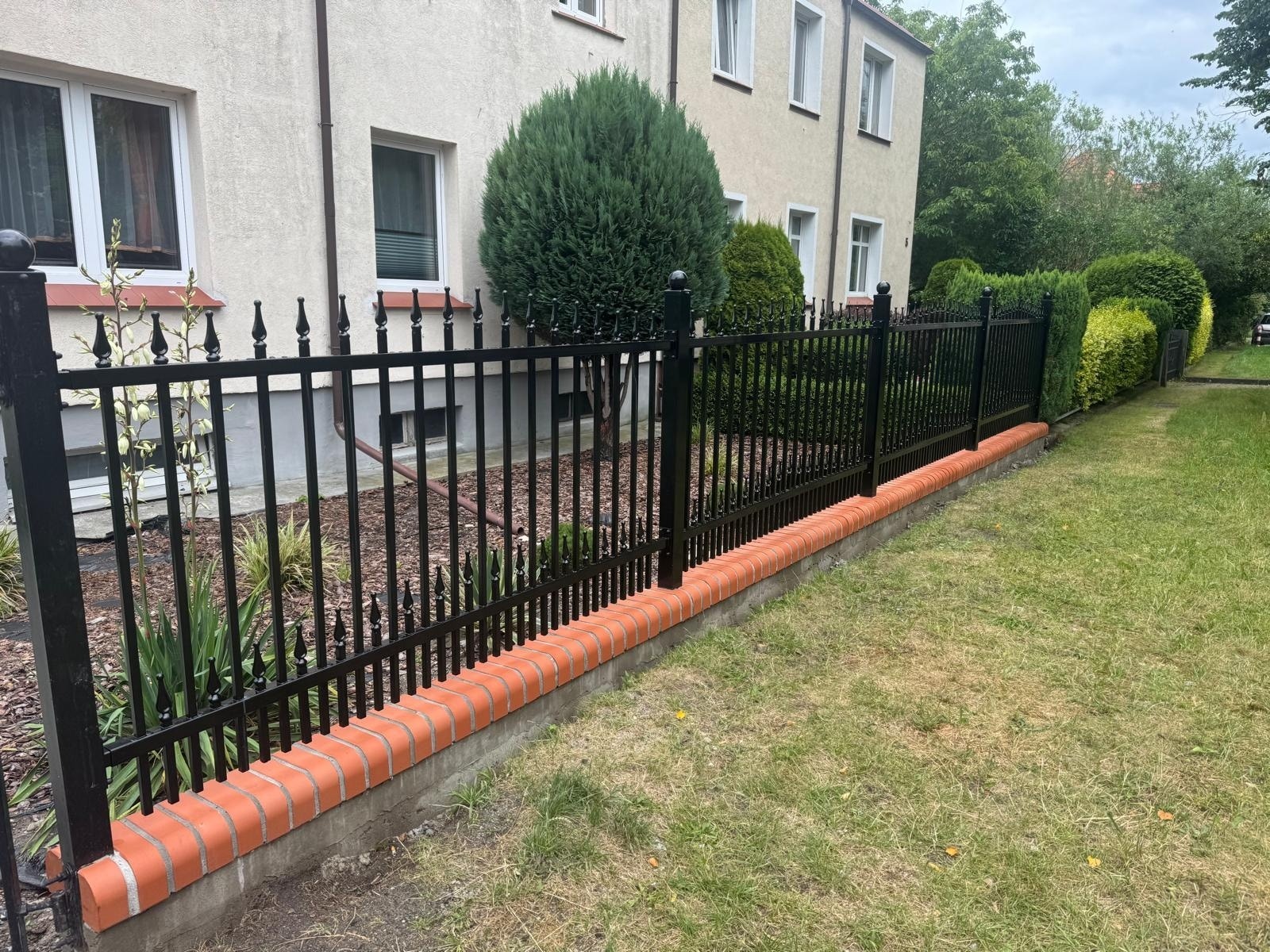 Eleganckie, czarne ogrodzenie metalowe z dekoracyjnymi grotami, osadzone na niskim murku z czerwonej cegły. W tle budynek i zieleń.