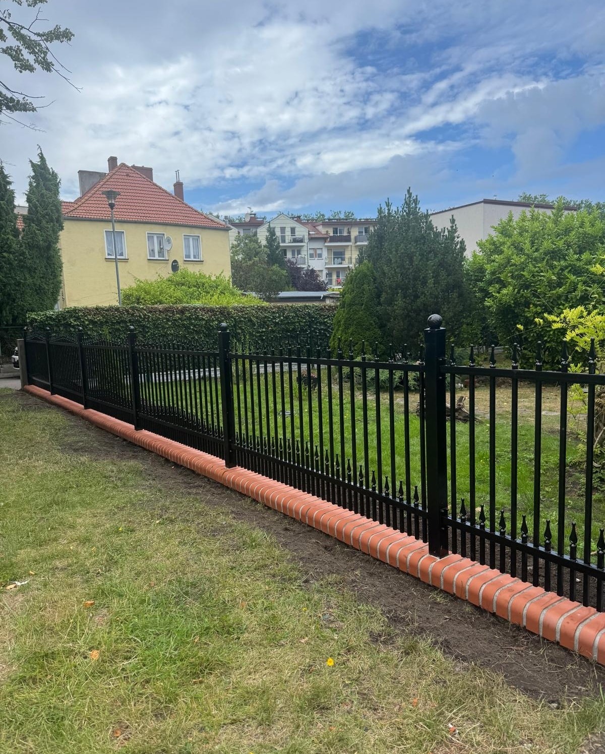 Czarne, metalowe ogrodzenie z ceglaną podmurówką na tle zielonego trawnika i zabudowy miejskiej w słoneczny dzień. Widoczne detale wykończenia.