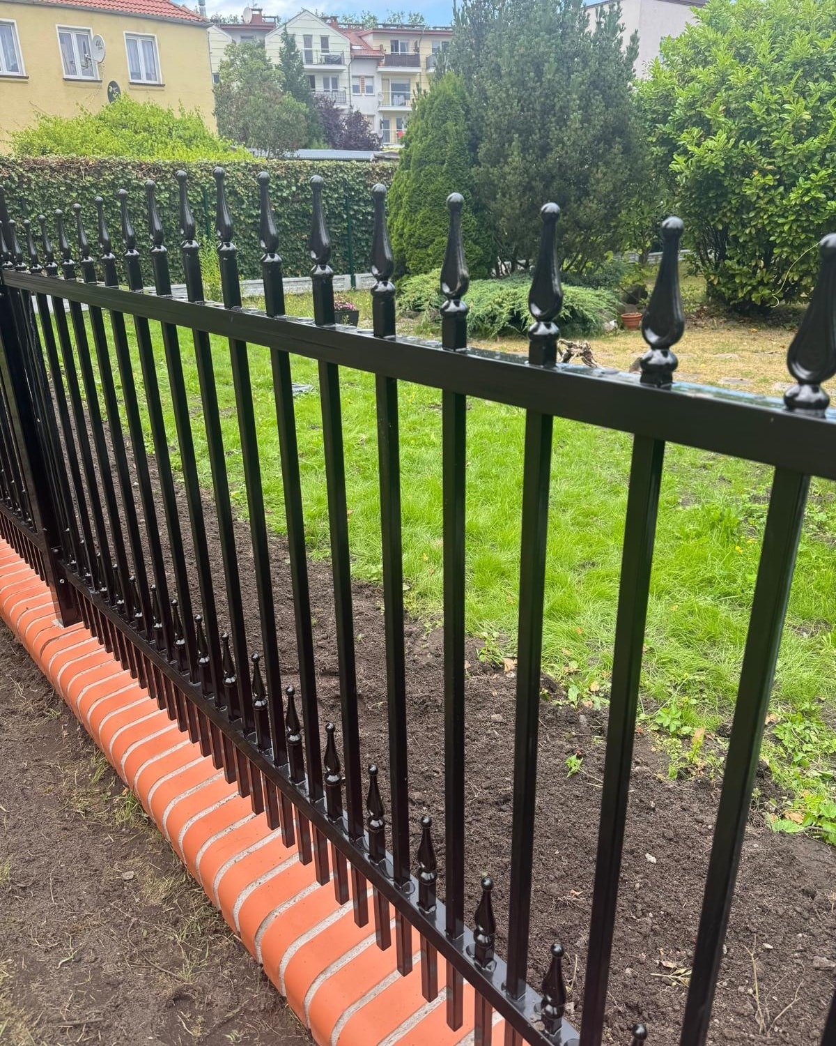 Czarne, metalowe ogrodzenie z dekoracyjnymi grotami, osadzone na niskim murku z czerwonej cegły. W tle zieleń trawnika i krzewów, budynki mieszkalne.
