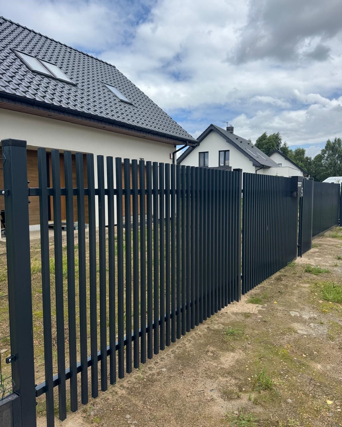 Nowoczesne, grafitowe ogrodzenie panelowe z pionowych profili, na tle domu z ciemną dachówką i pochmurnego nieba. Widoczne słupki i brama wjazdowa.